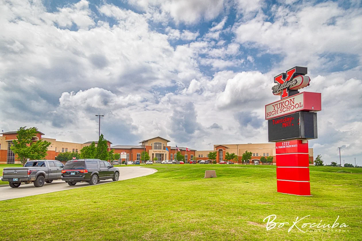 Yukon High School in Yukon, Oklahoma, featuring the large red school entrance sign, landscaped lawn, and school buildings beneath a partly cloudy sky.