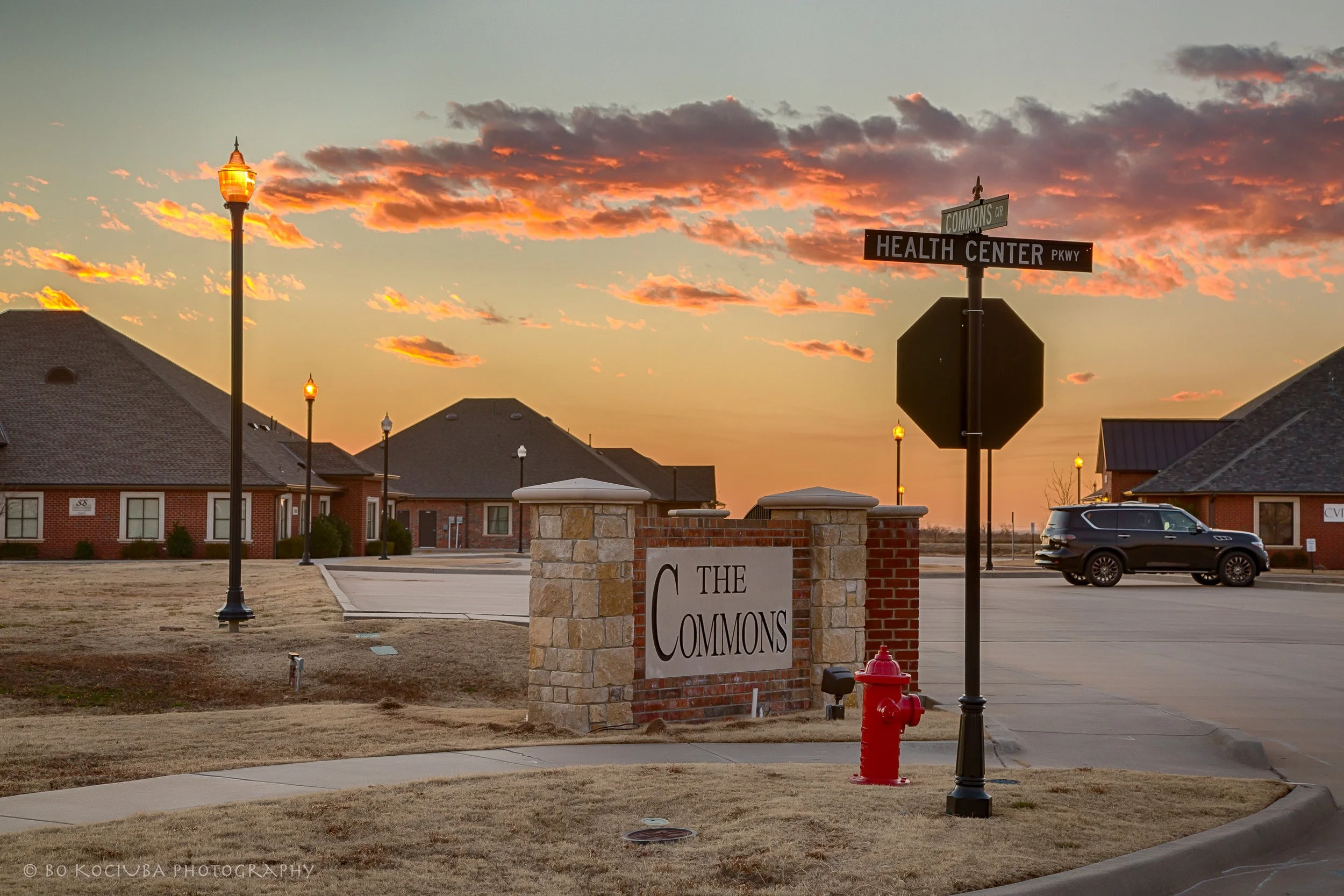 The Commons commercial area near Garth Brooks Boulevard in Yukon, Oklahoma, at sunset, showing bricked business structures and the intersection of The Commons Circle and Health Center Parkway.