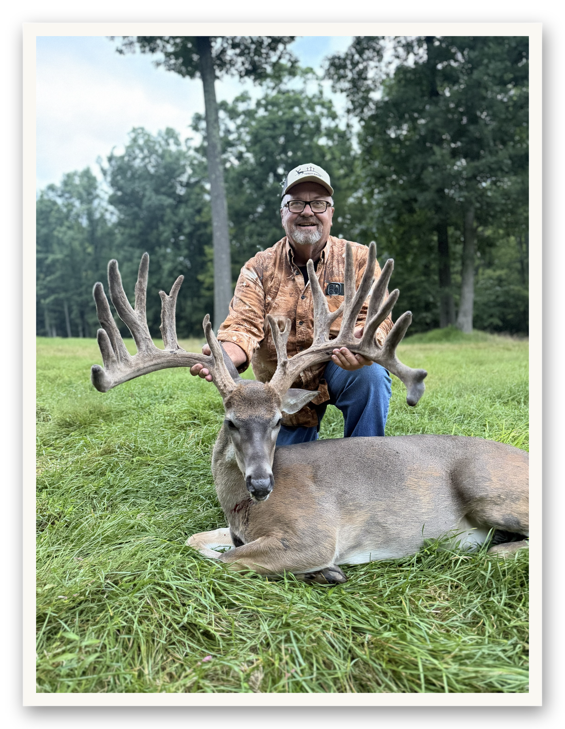 Original phone photo of a hunter kneeling behind a record-class whitetail buck in velvet in a green field.