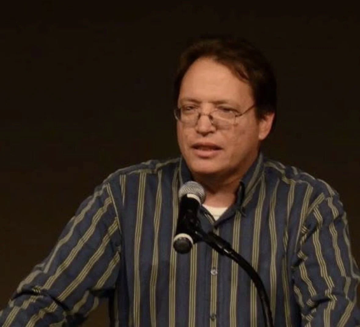 A man with glasses wearing a striped shirt speaking into a microphone.