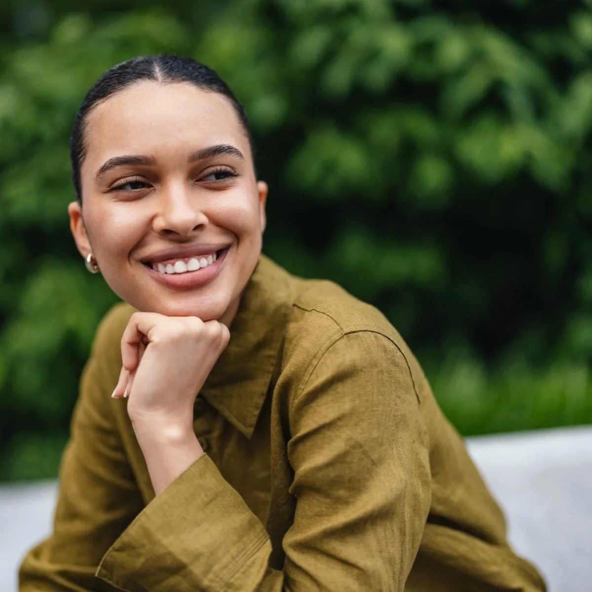 A young woman with dark hair pulled back, smiling and looking to the side, resting her chin on her hand. She is wearing a mustard-colored jacket and a small earring, with a blurred green outdoor background.