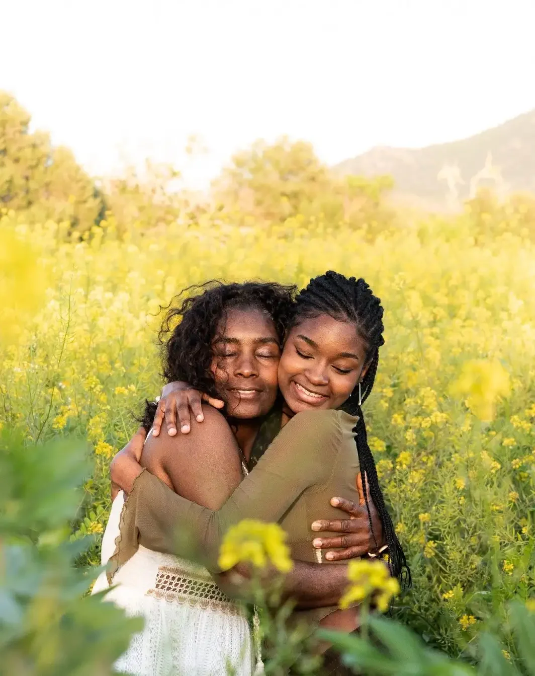 Two women hugging each other and smiling in a yellow flower field with trees and mountains in the background.