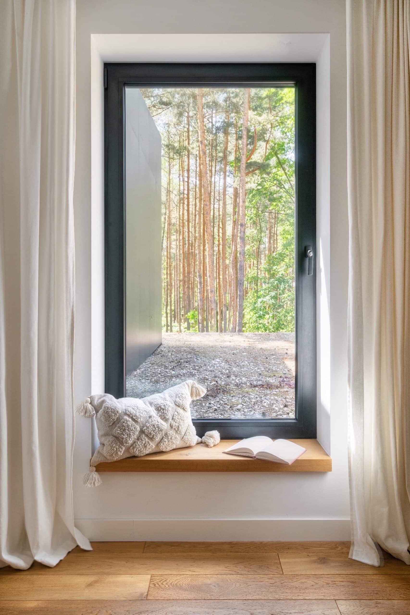 View through a window showing tall trees and green foliage outside, with cream curtains on either side and a window seat with a white textured cushion and an open book inside.