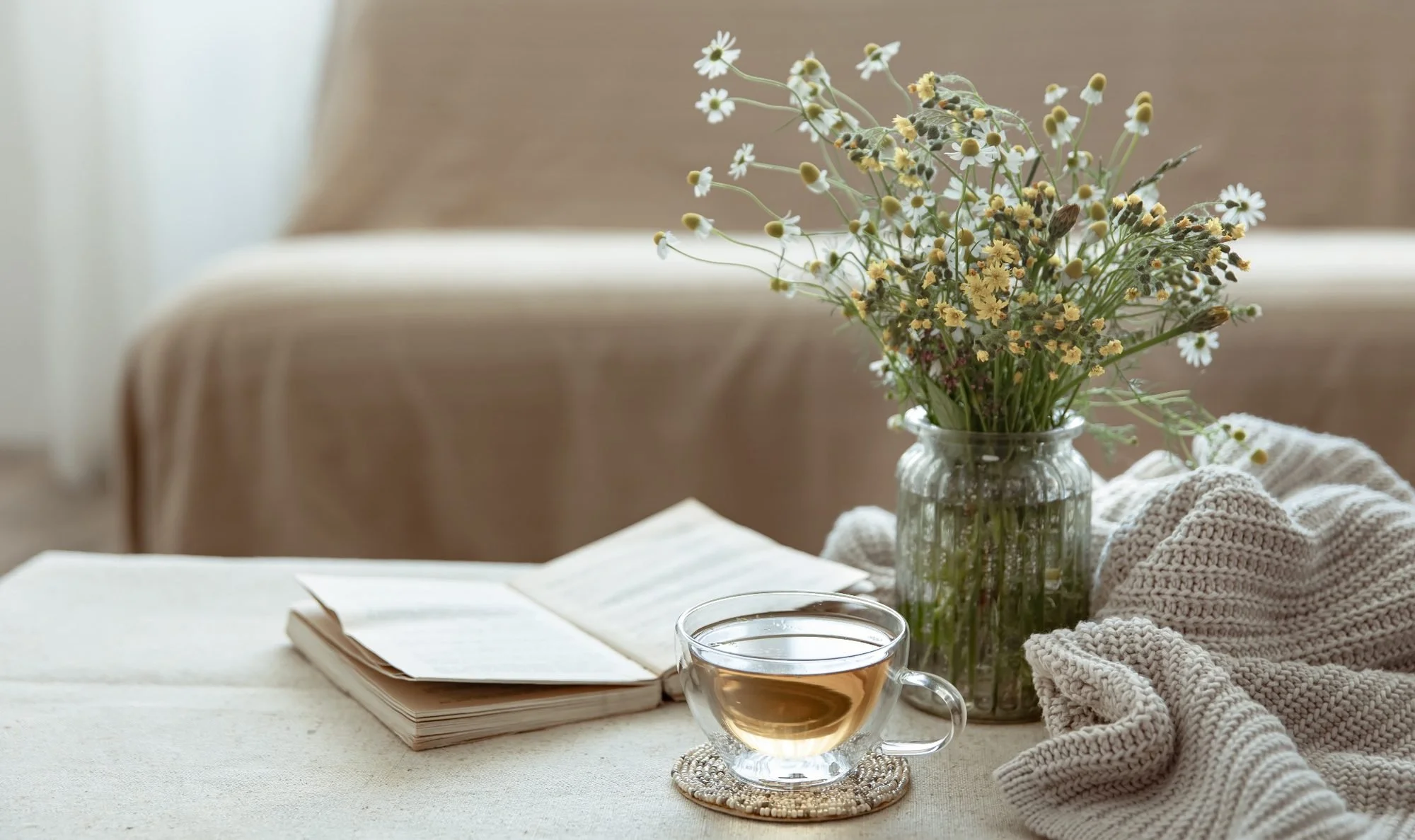 A table with a bouquet of flowers in a glass vase, an open book, a glass cup of tea, and a cozy knitted blanket.