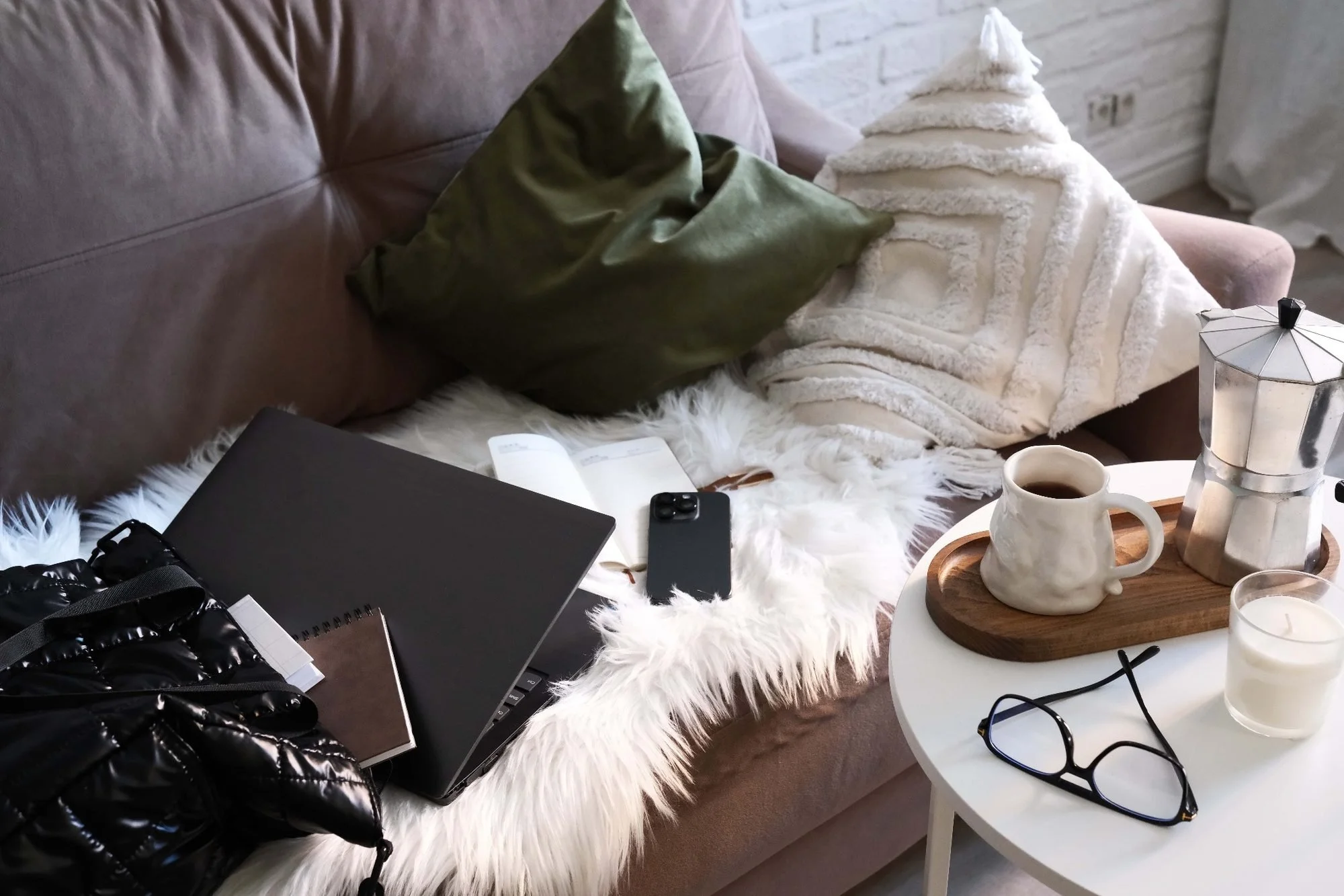 Cozy living room with a brown sofa, green pillow, and a fluffy white blanket. On the coffee table, there's a cup of coffee, a moka pot, a notebook, glasses, and a smartphone. The space is decorated with a white fur throw and a white brick wall.