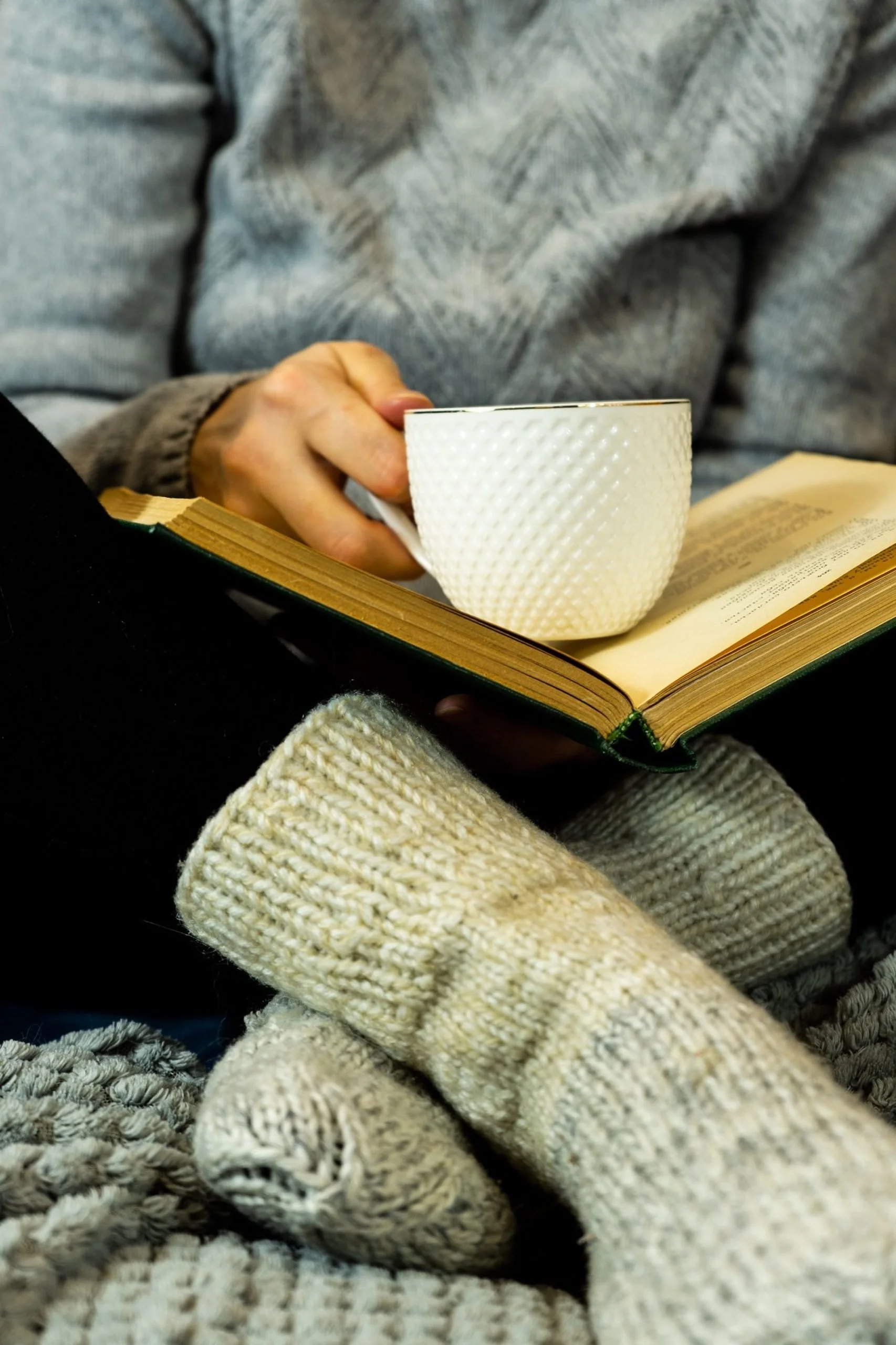 Person wearing a gray sweater holding a white textured cup above an open book with glasses and cozy beige and gray blankets nearby.