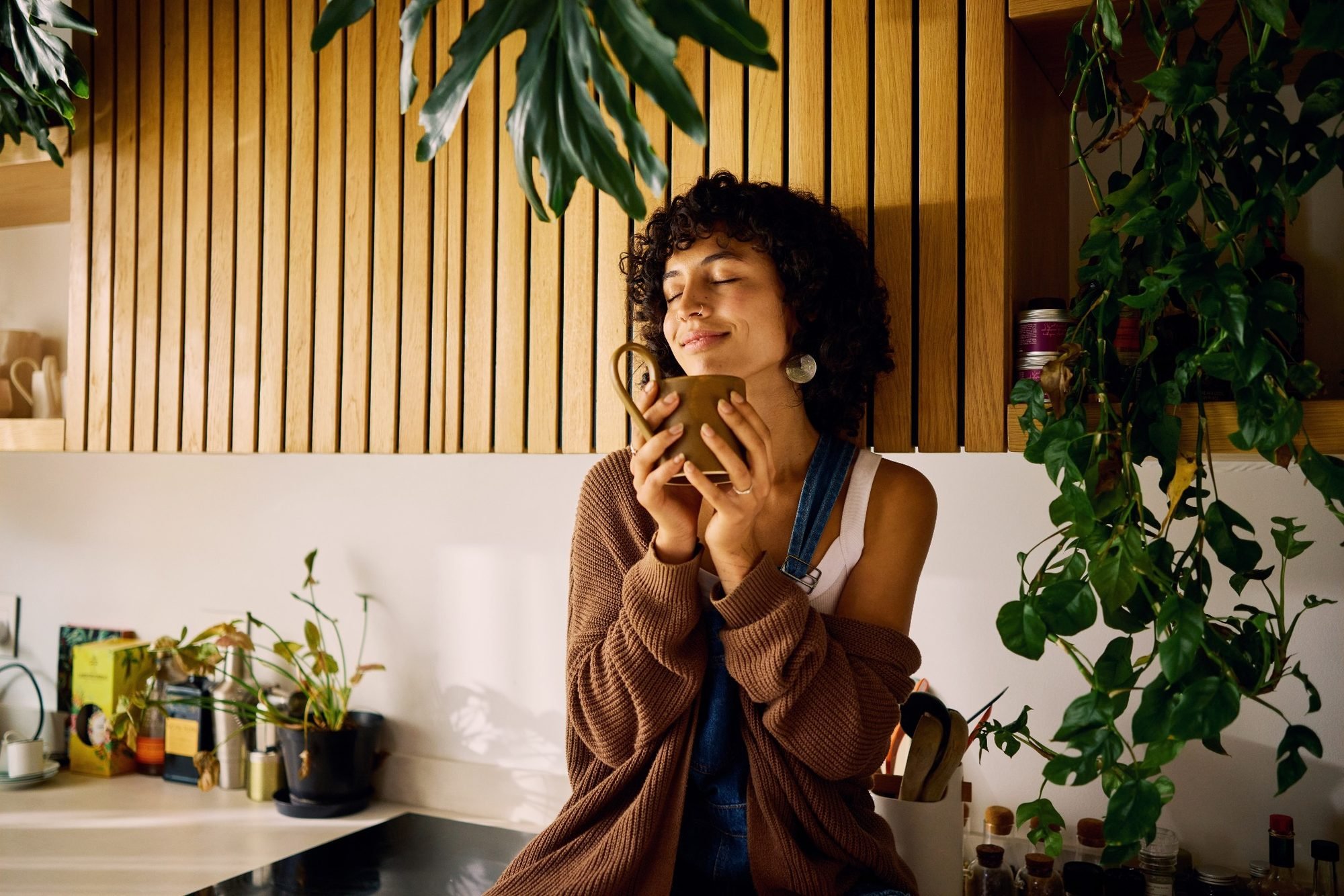 A woman with dark curly hair and a nose ring, smiling with eyes closed, holding a tan mug, standing in a cozy indoor space with wooden paneling and green plants.