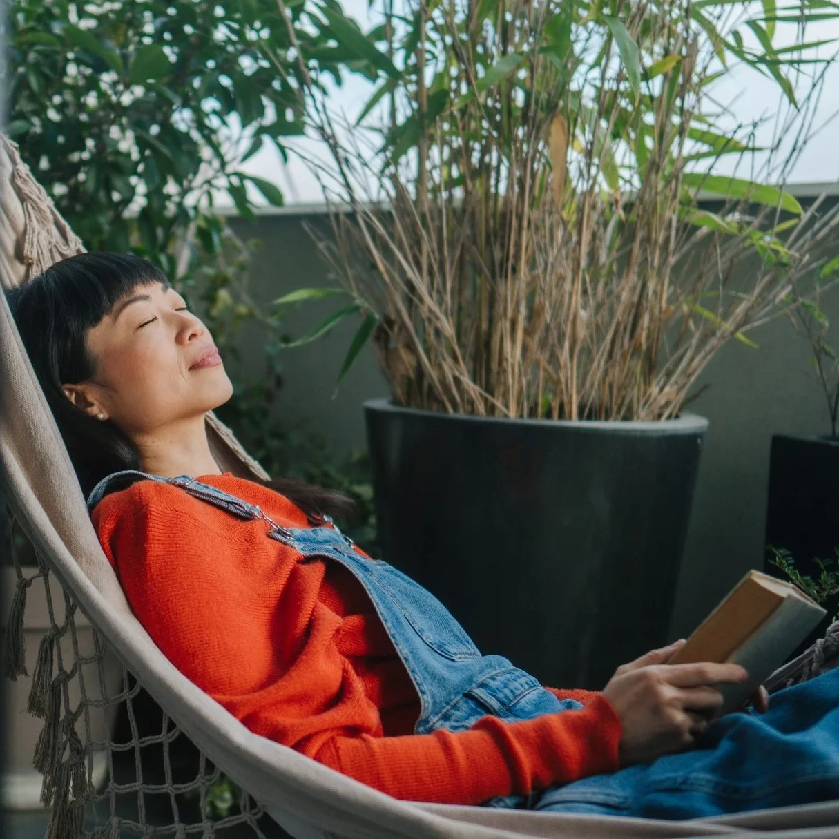 A woman with short black hair and bangs relaxing in a hammock, holding a book, surrounded by green plants on a balcony.