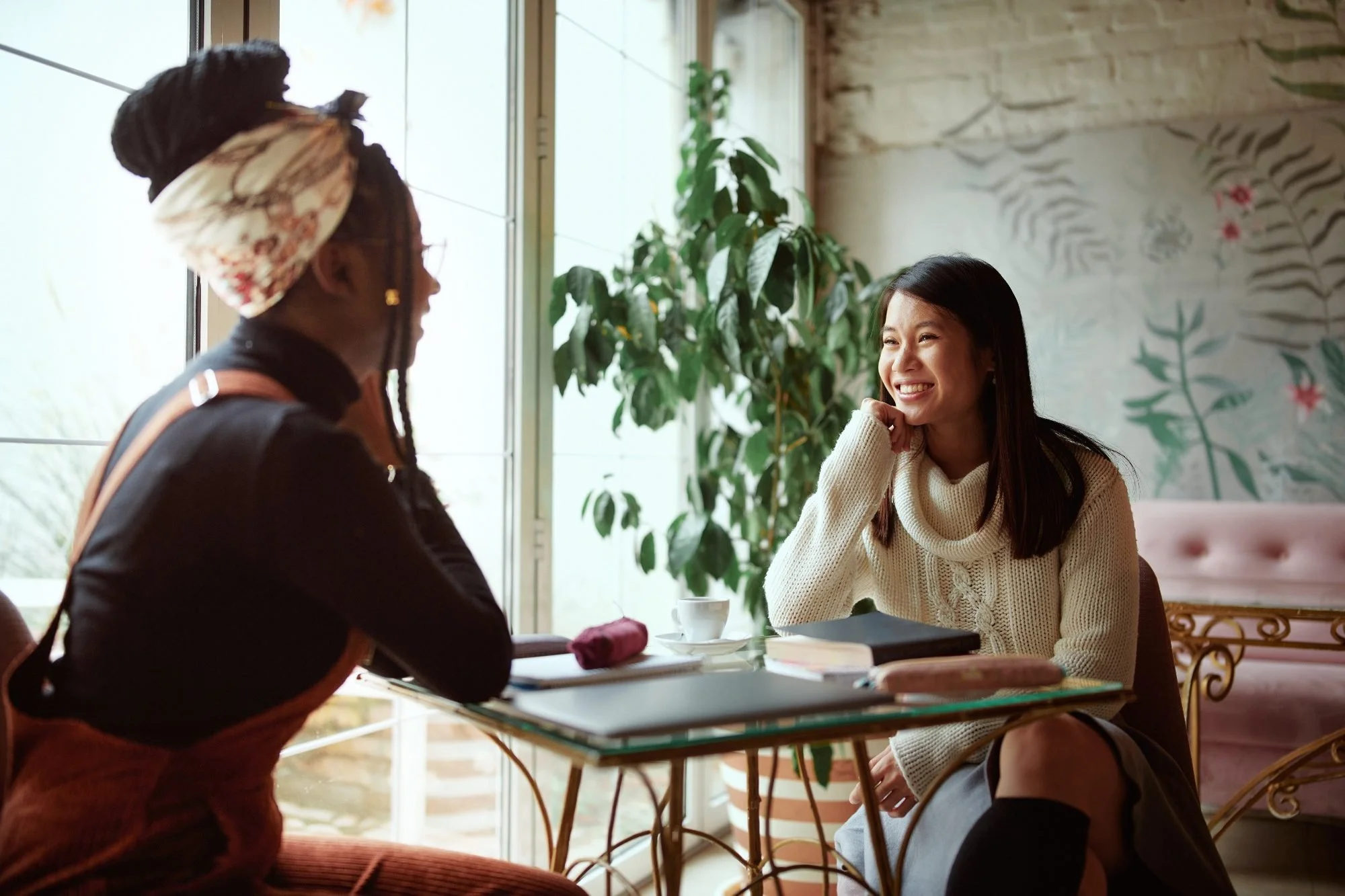 Two women at a cafe, one with long dark hair smiling and resting her chin on her hand, the other with braided hair in a headscarf, seen in profile, seated at a table with a coffee cup, books, and a purple cloth, with green plants and a wall mural in the background.