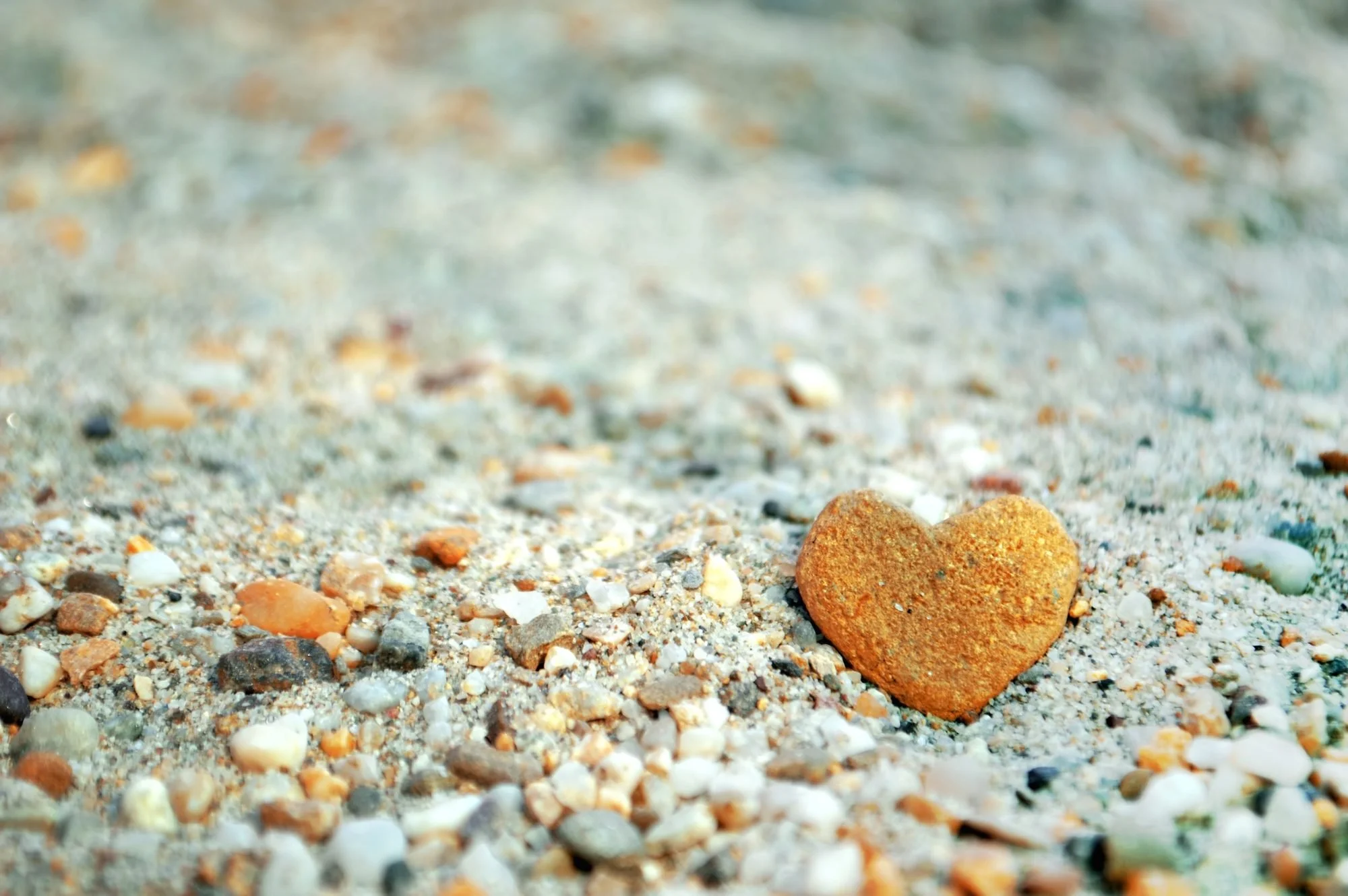 A close-up of a sandy beach with small colorful pebbles and a single heart-shaped rock in the foreground.