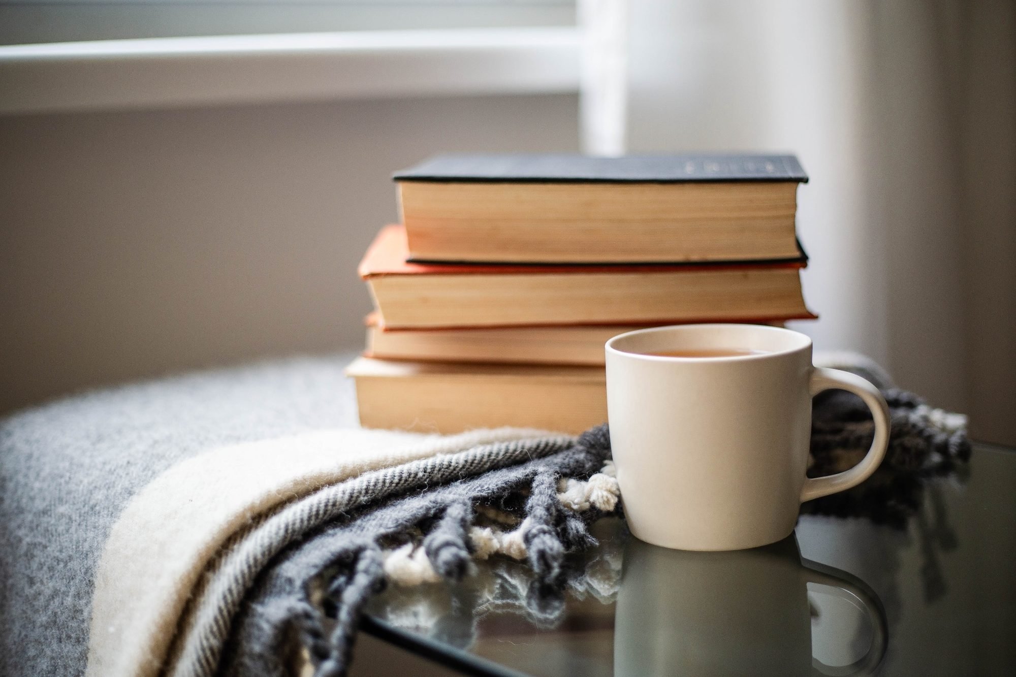 A stack of five hardcover books with different colored covers sitting on a gray surface near a window, with a white cup of coffee or tea nearby, partially on a patterned black and white cloth.