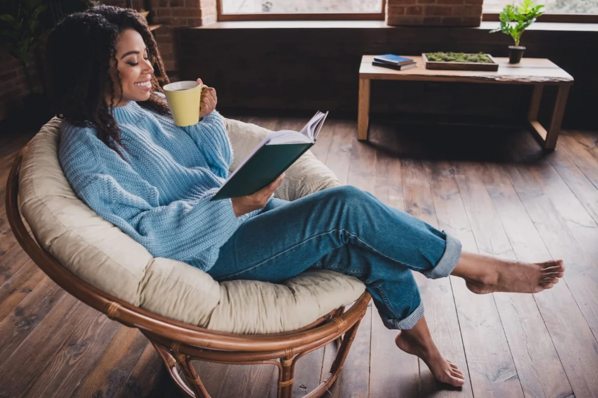 A woman with curly hair is sitting on a round cushioned chair, holding a book in one hand and a yellow mug in the other, smiling while reading. She is wearing a blue sweater and jeans, with one leg crossed over the other. The room has wooden flooring, a window, and a wooden table with plants and books in the background.