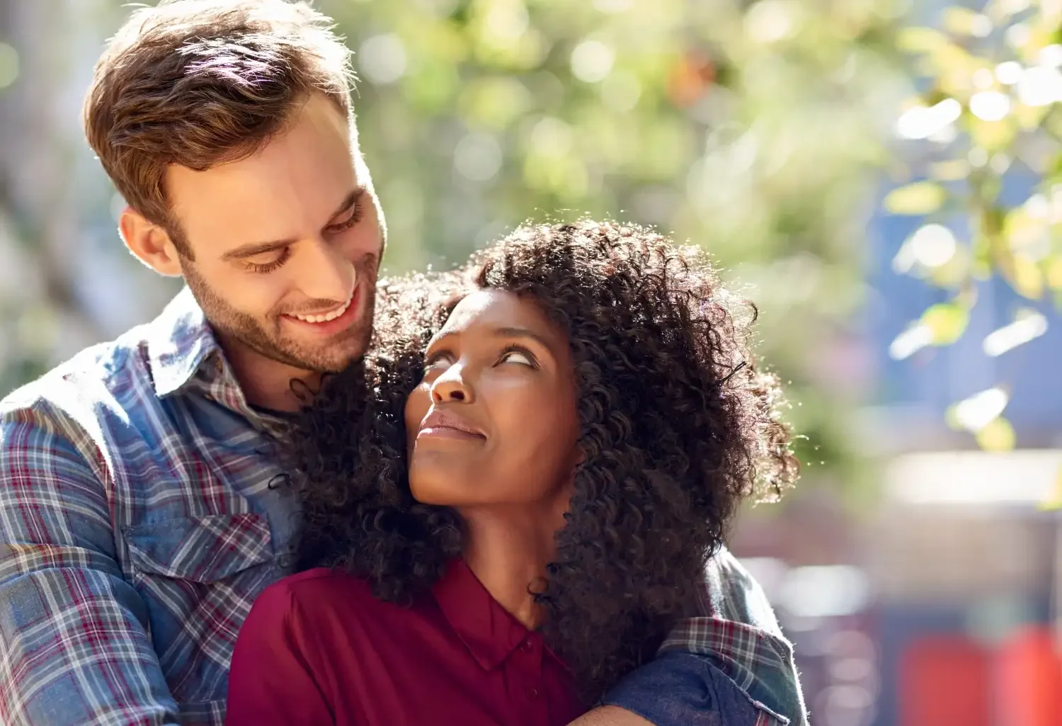 A man and a woman sharing a warm embrace outdoors, with sunlight filtering through trees in the background. The man is smiling down at the woman, who is looking up at him with a smile.