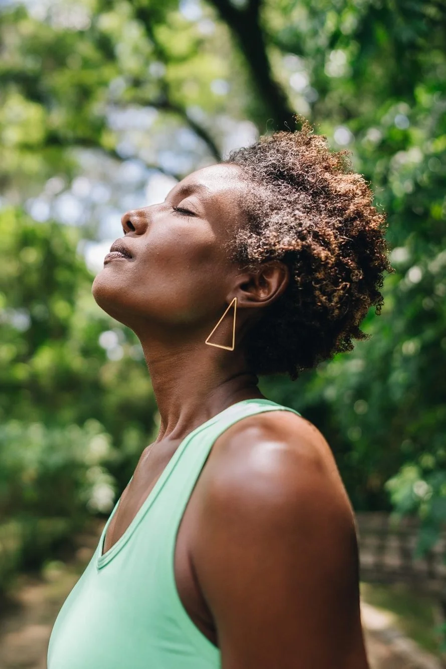 A woman with short curly hair in a natural outdoor setting, eyes closed, wearing a light green sleeveless top and triangular gold earrings.