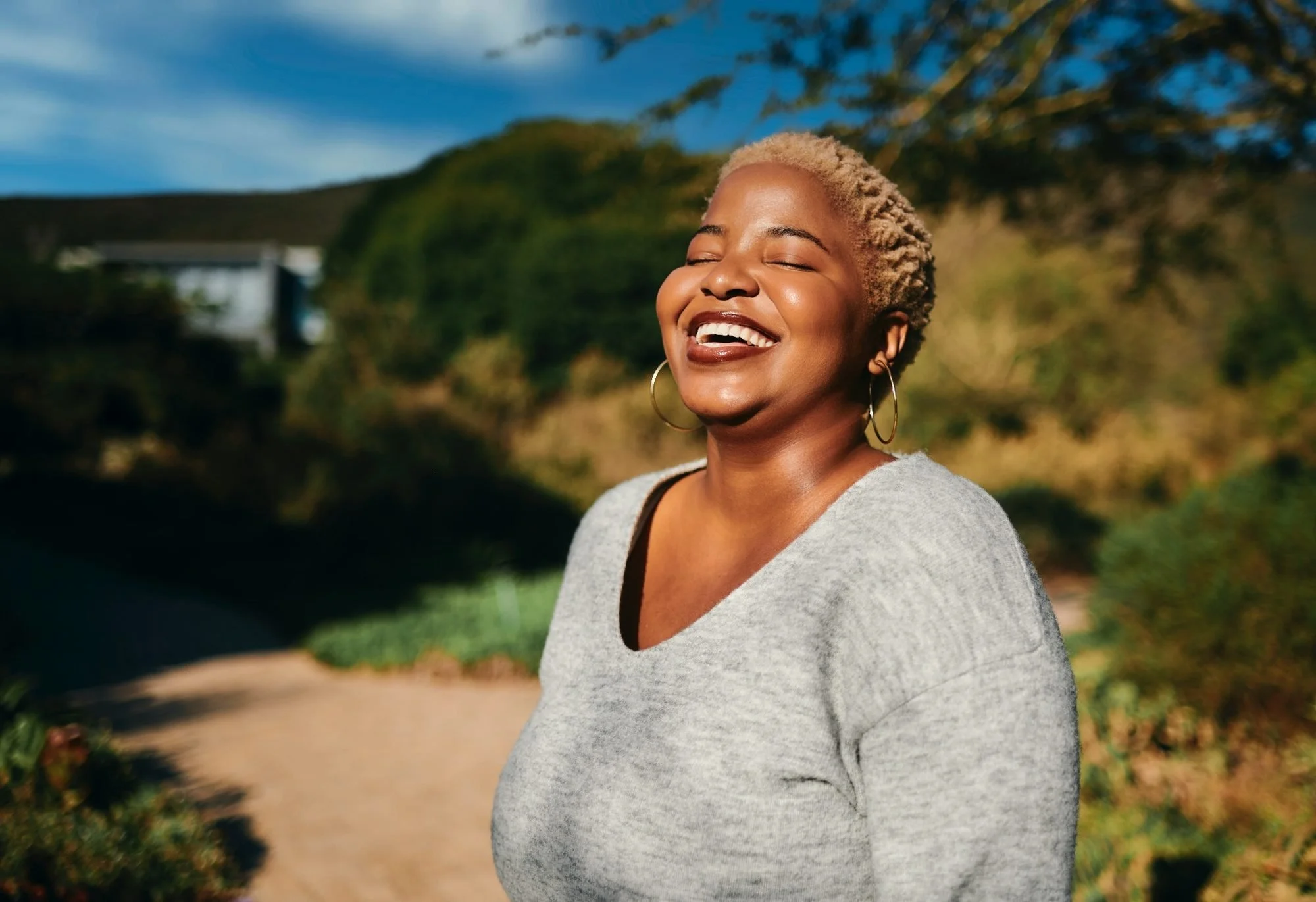 A smiling woman with short, blonde hair and hoop earrings standing outdoors in a park with trees and a building in the background.