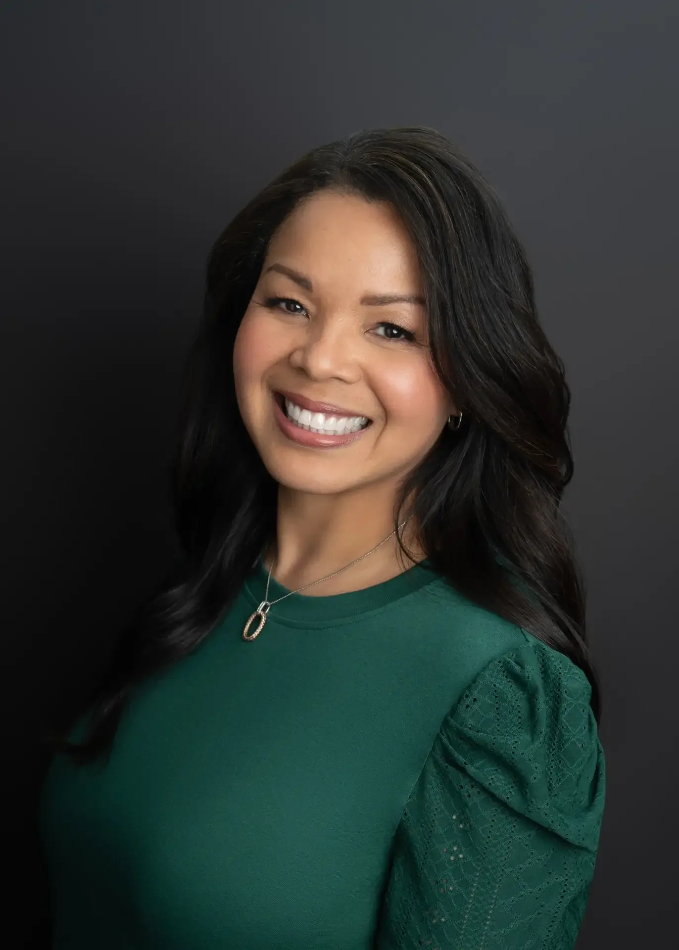Portrait of a woman smiling, wearing a green top with lace detailing on the sleeves, a necklace with a pendant, and earrings, against a dark background.