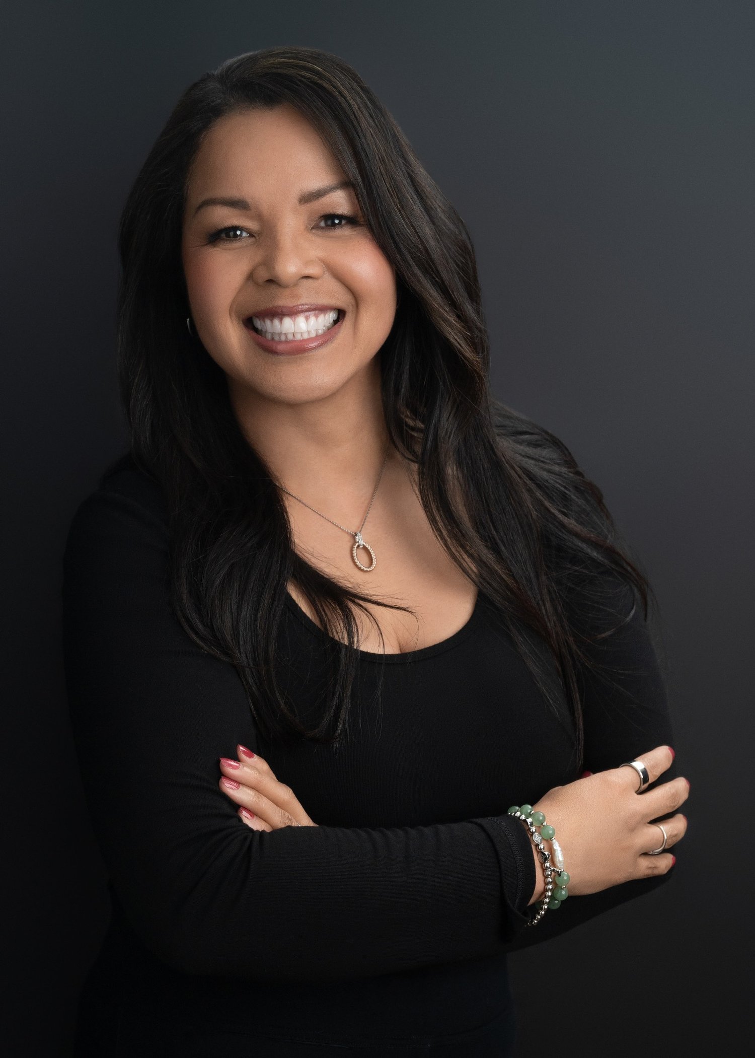 Close-up of a woman with long dark hair smiling against a dark background, wearing a black top and jewelry.