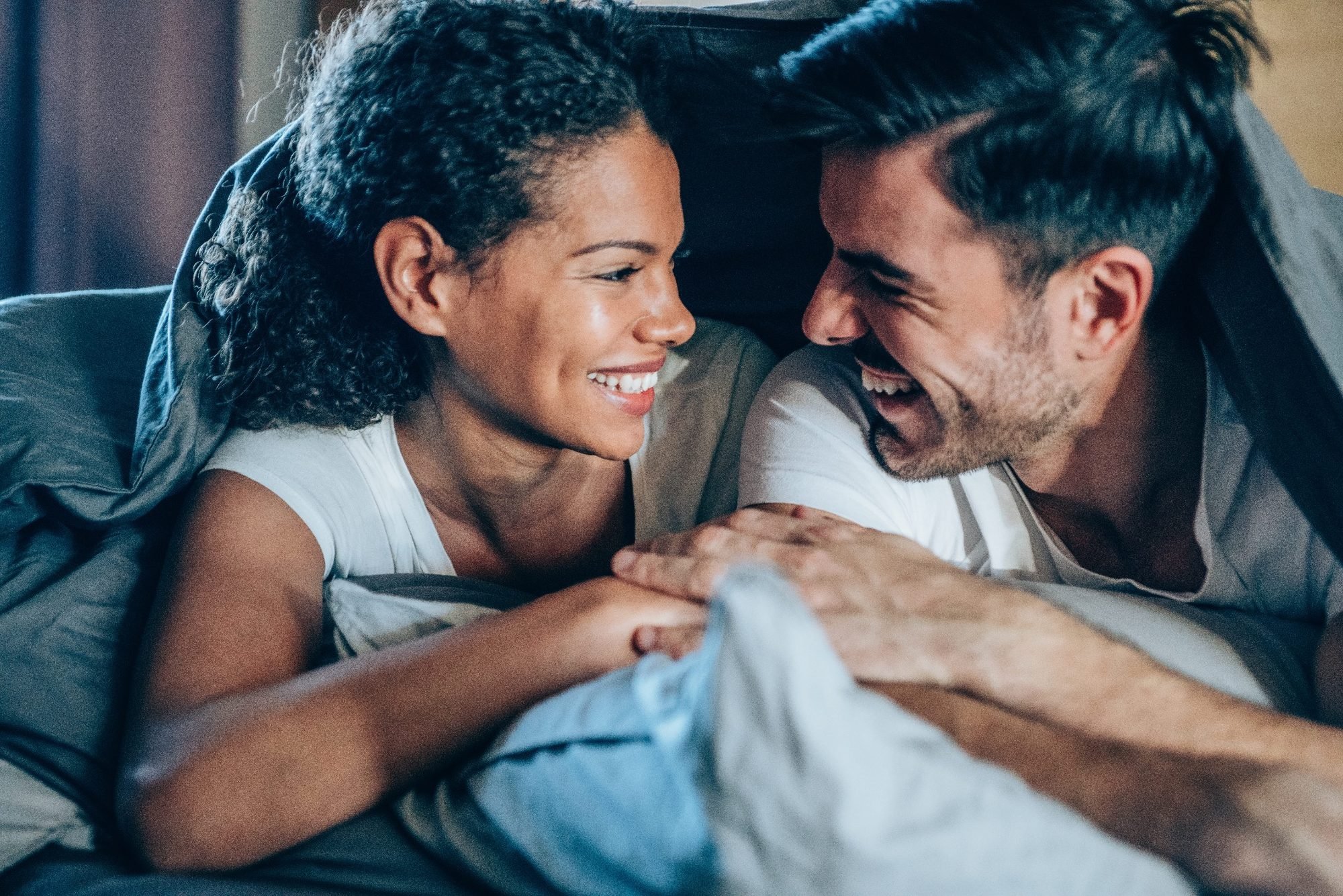 A woman and a man are smiling and sitting close together on a bed, leaning their foreheads against each other, appearing happy and affectionate.