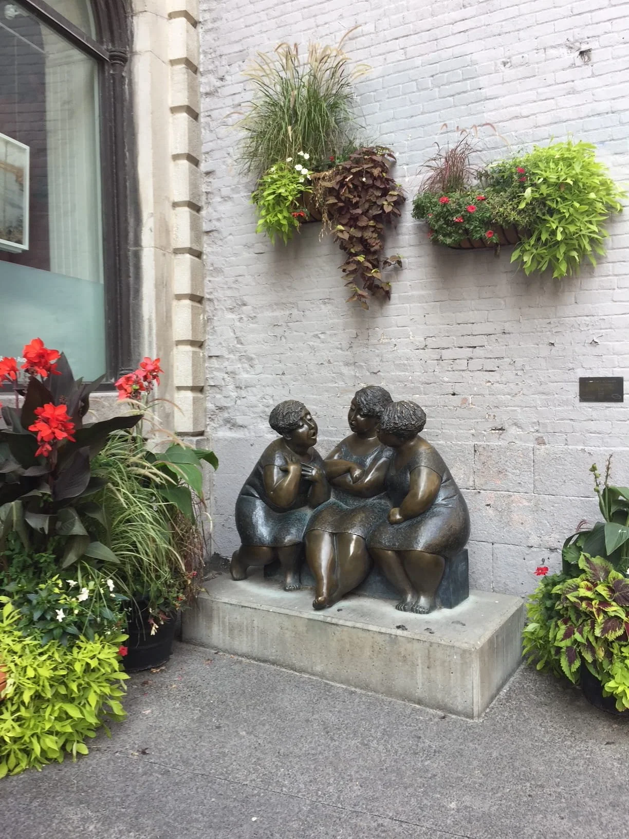 Bronze sculpture of three women sitting together, engaged in conversation, located outdoors against a white brick wall decorated with hanging flower baskets and surrounded by potted plants.