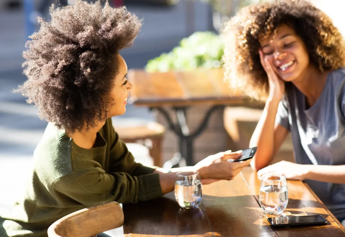 Two women with curly hair sitting at a wooden table, smiling, with glasses of water and a smartphone in hand, outdoors on a sunny day.