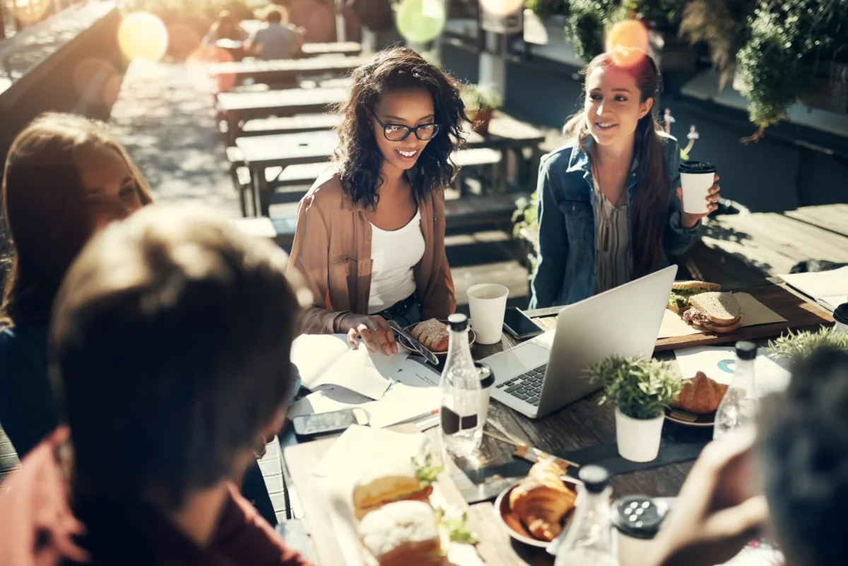 Group of friends having a meal outside at a wooden table, smiling and talking, with food, drinks, a laptop, and notebooks in front of them, in a sunny outdoor setting.