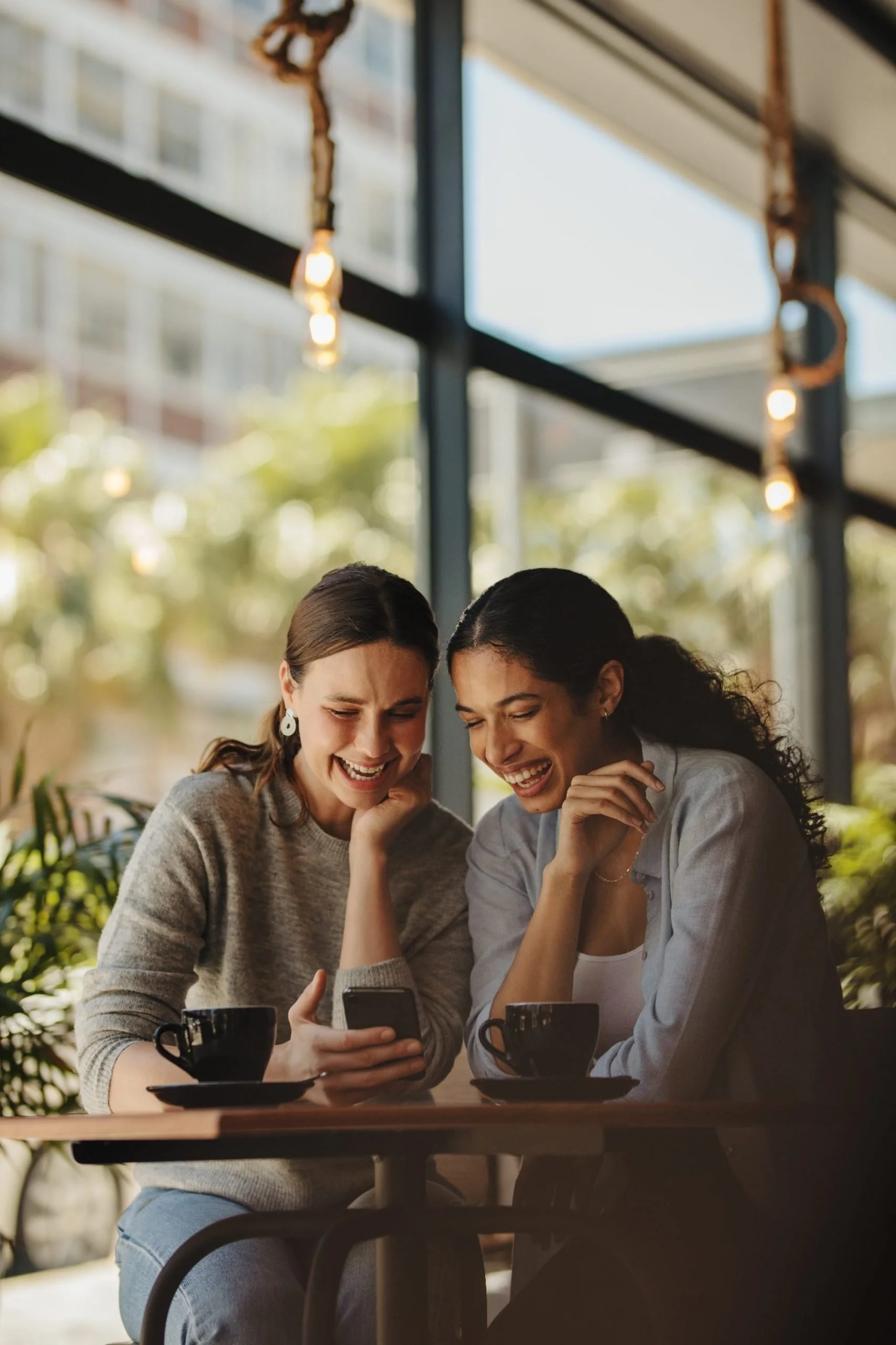 Two women are sitting at a table in a cafe, looking at a smartphone, smiling, with coffee cups in front of them, inside a brightly lit space with large windows and hanging lights.