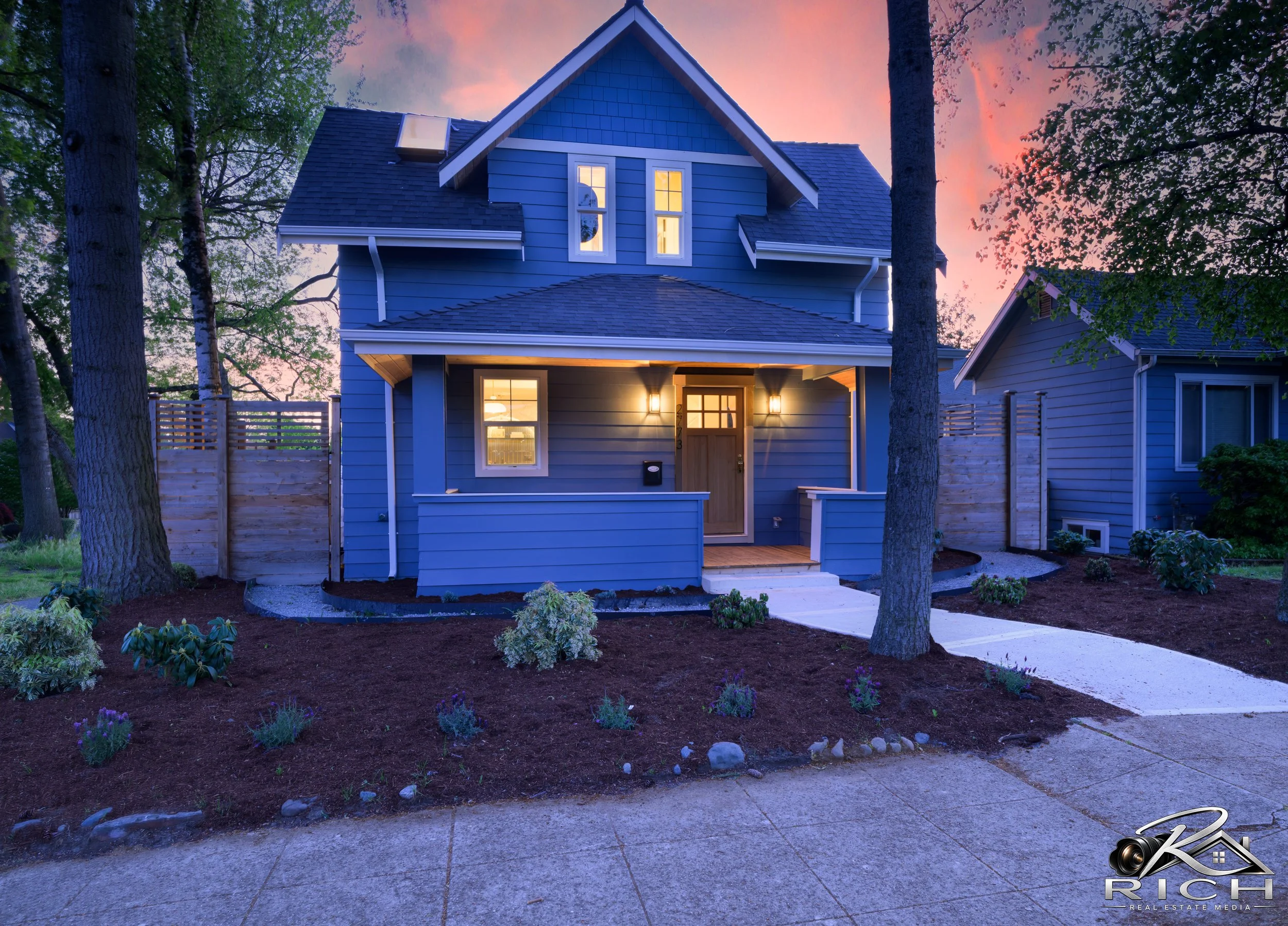 A blue house with white trim and a gabled roof, illuminated by exterior lights at dusk, surrounded by trees and a well-maintained garden with small plants and a concrete pathway leading to the front door.