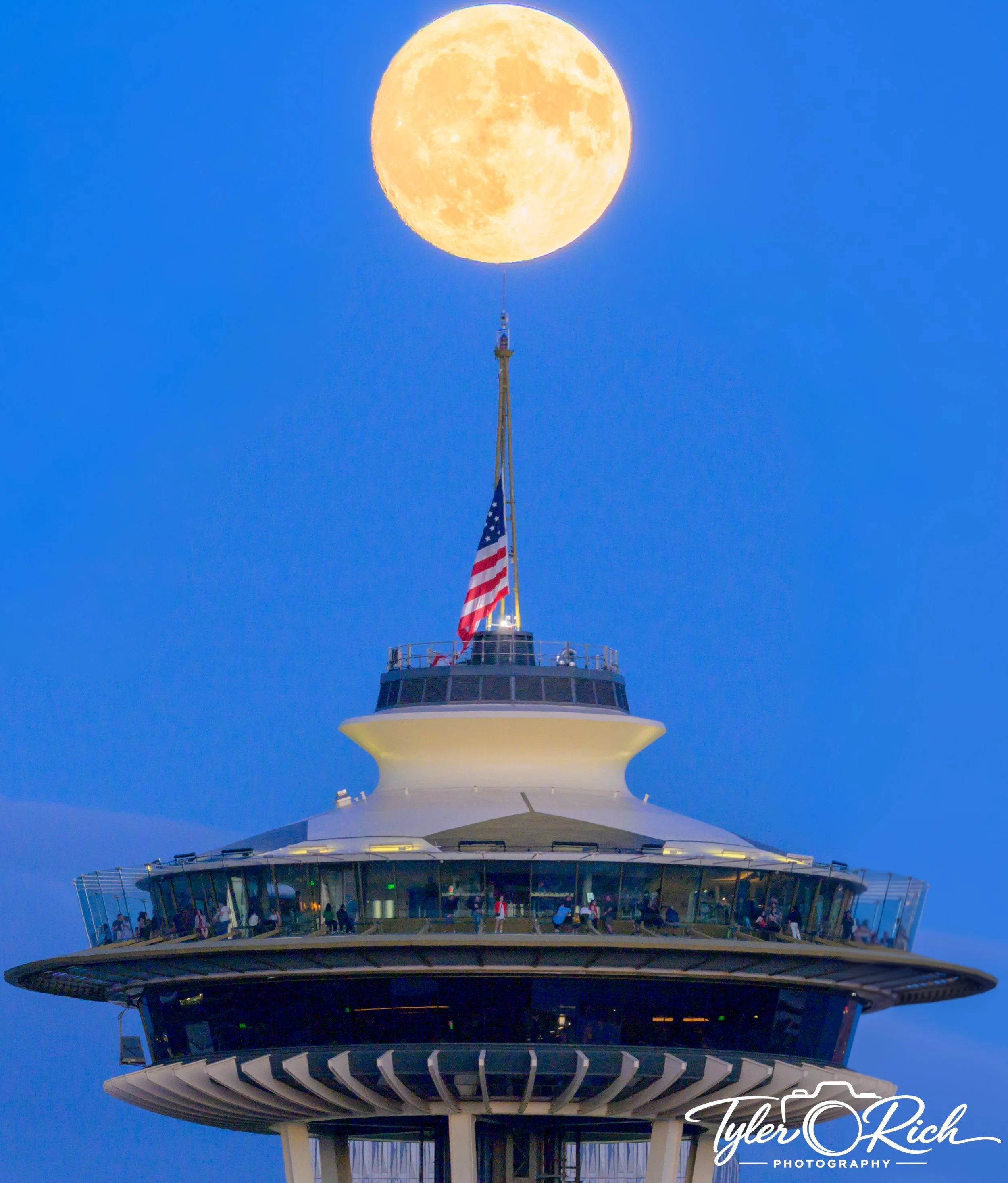 The Space Needle in Seattle at dusk with a full moon rising behind it and an American flag flying near the top.