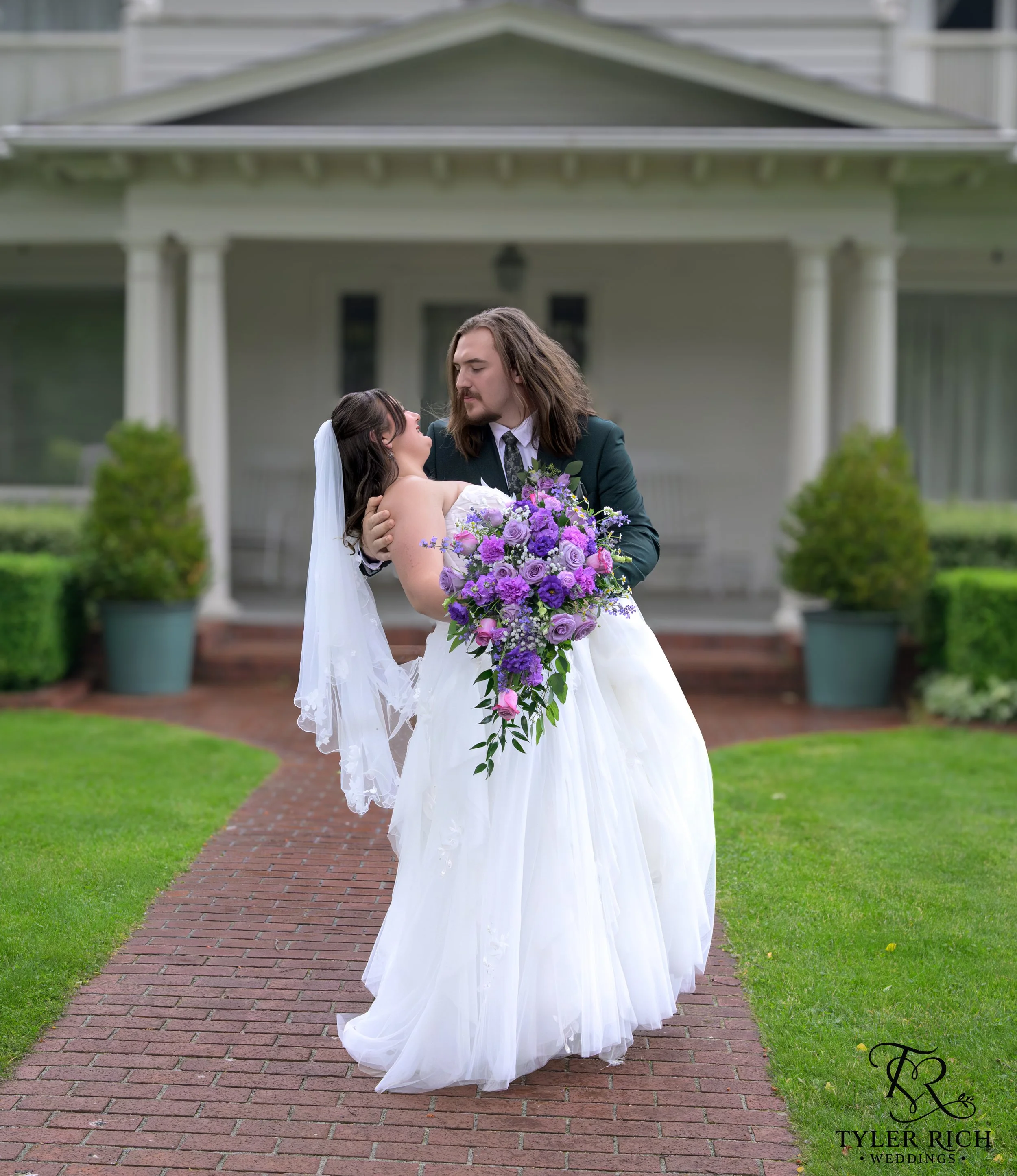 A bride and groom with long hair stand closely together on a brick pathway in front of a house with white columns. The bride holds a large bouquet of purple and pink flowers, and the groom is wearing a dark suit. They are gazing into each other in a romantic moment.