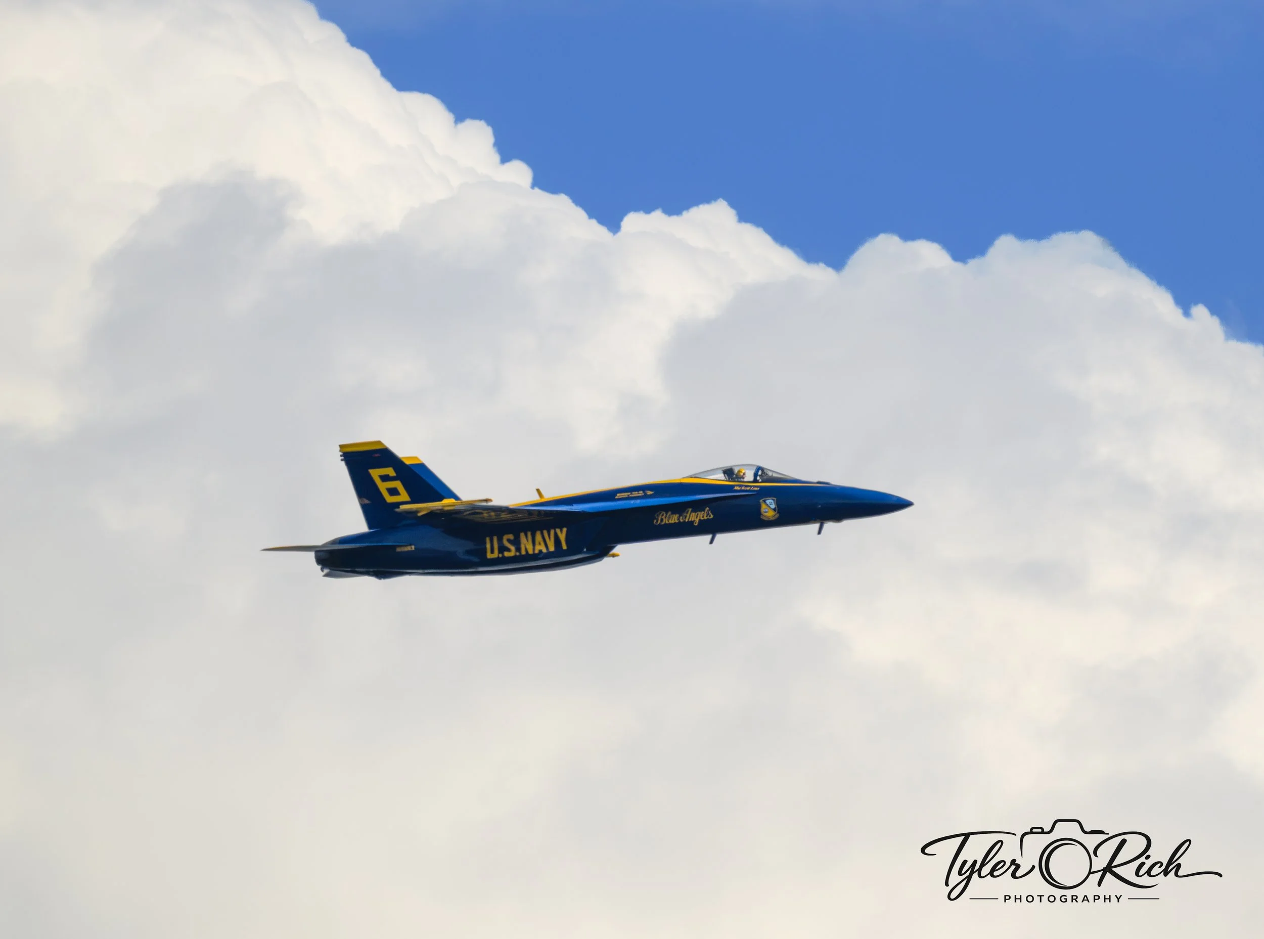 A blue and yellow U.S. Navy Blue Angels jet aircraft flying in the sky among white clouds.