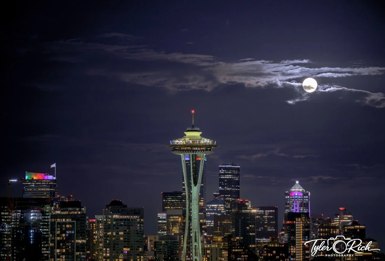 Nighttime view of Seattle skyline with illuminated city buildings, the Space Needle in center, and a full moon partially covered by clouds in dark sky.