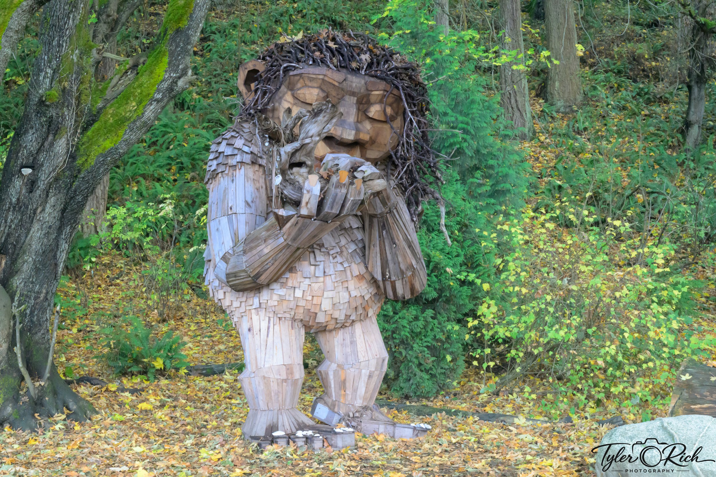 Wooden sculpture of a person with long hair, eyes closed, holding their hands to their face, situated outdoors among trees and green foliage.