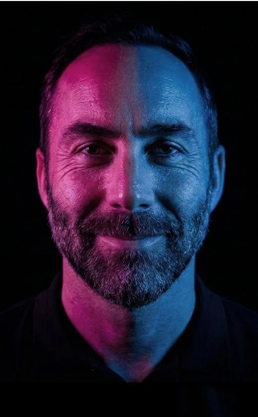 A man with a beard smiling, illuminated with pink and blue lighting against a black background.