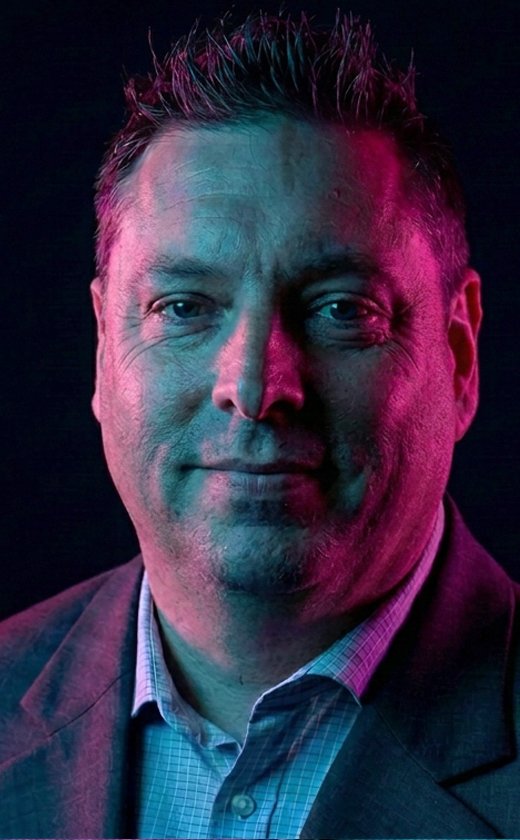 Portrait of Patrick Smith, creative director, wearing a blazer and collared shirt, illuminated by blue and pink lighting against a dark background.