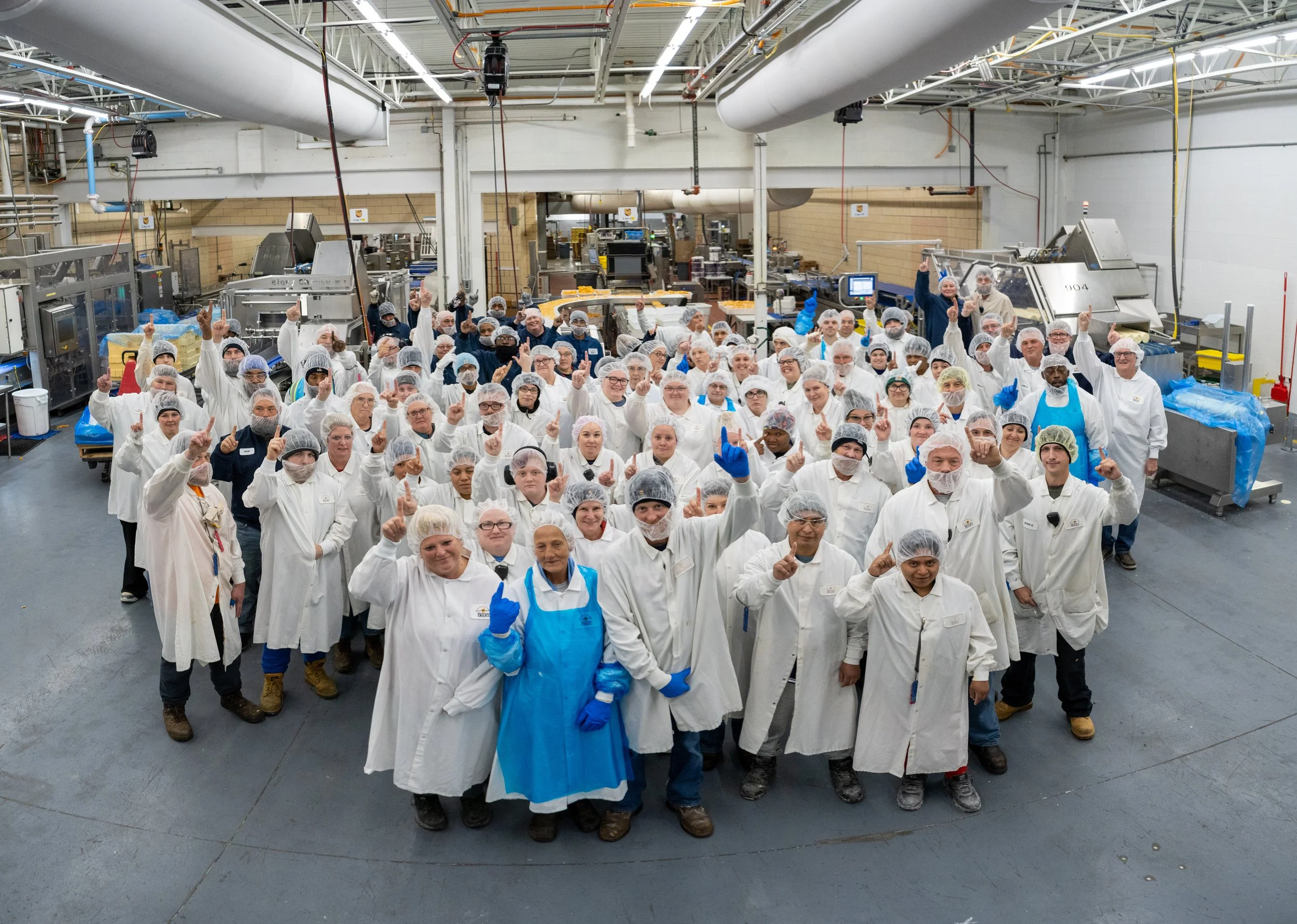 A large group of workers in white lab coats, hairnets, and gloves gathered together in a factory or processing plant. They are posing for a group photo, with many raising their index fingers. The factory has industrial equipment and machinery in the background.