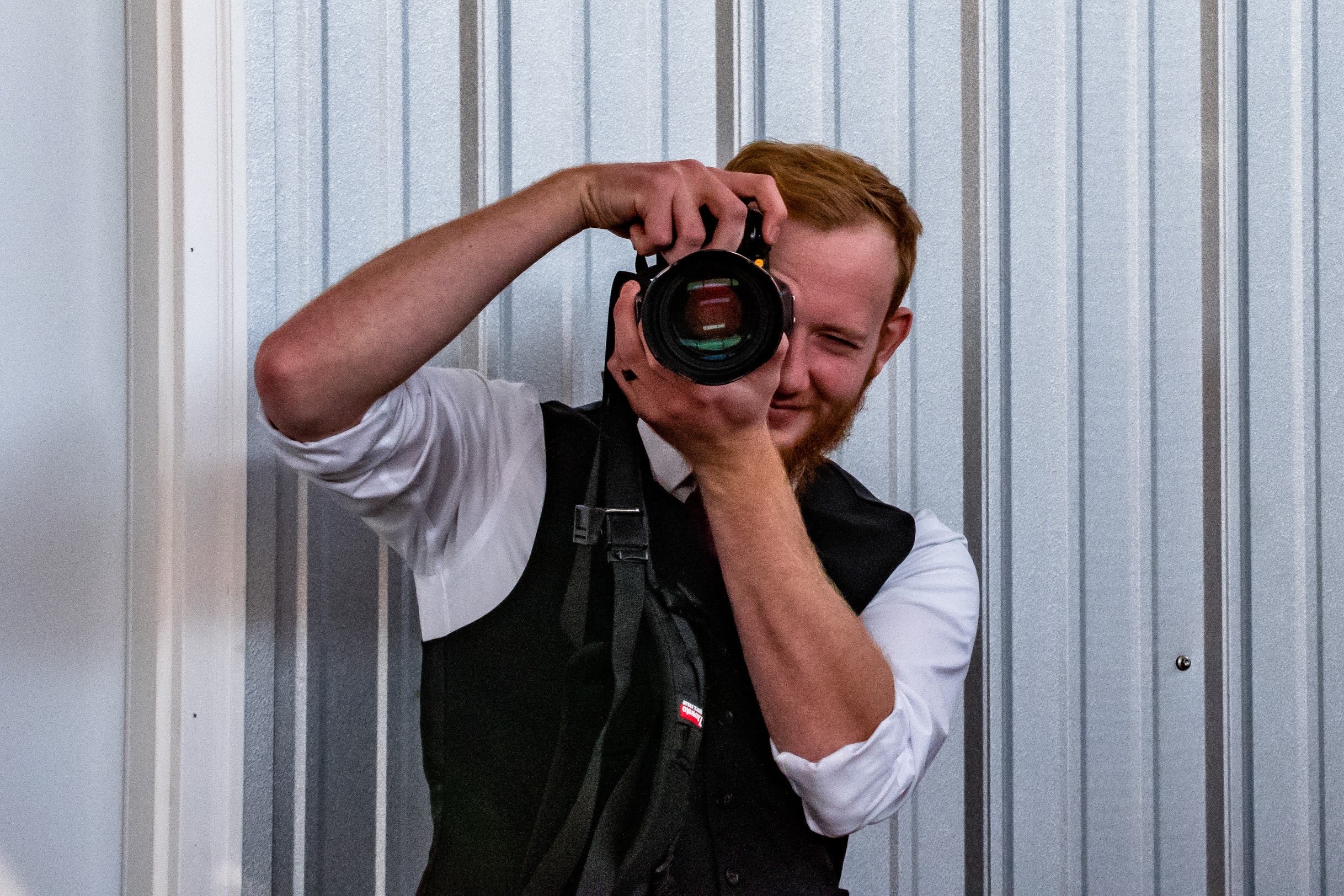 A man with red hair and a white shirt taking a photo of himself in a mirror, with a striped wall in the background.