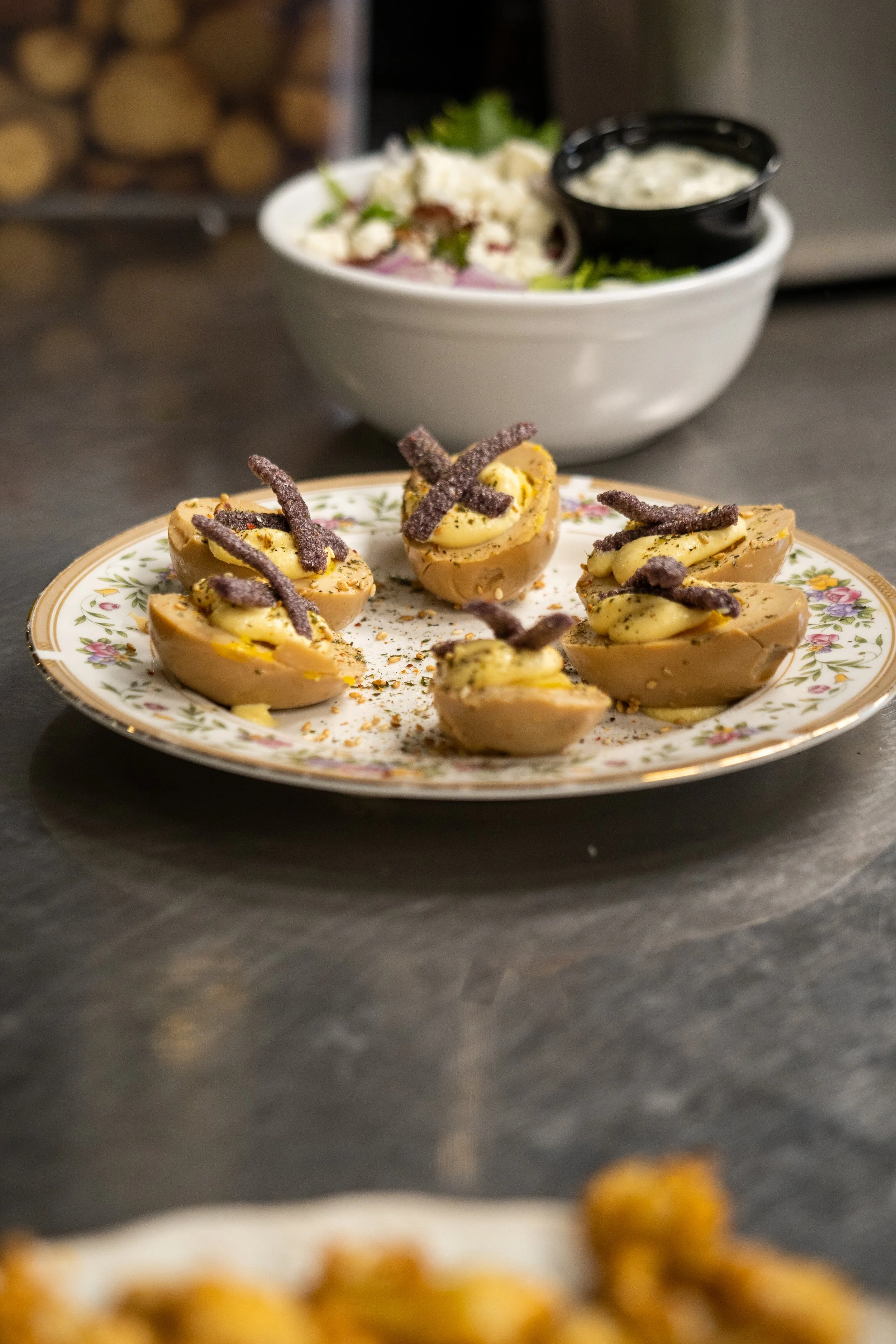 A plate with deviled eggs garnished with bacon strips and herbs, with a bowl of potato salad and another dish in the background.