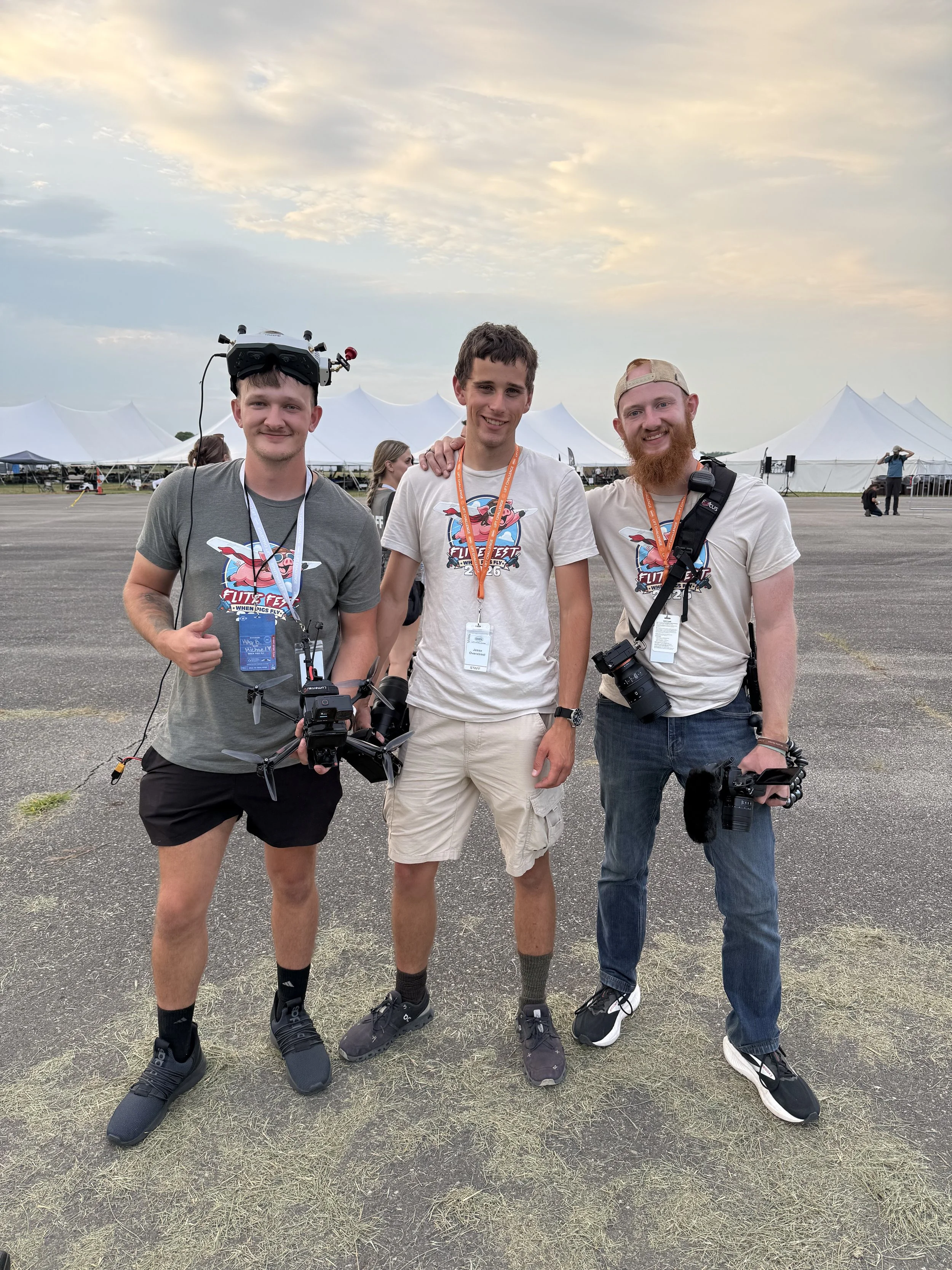 Three young men standing outdoors in front of white event tents, wearing casual clothes and event badges, with drones and camera gear, smiling at the camera during sunset.