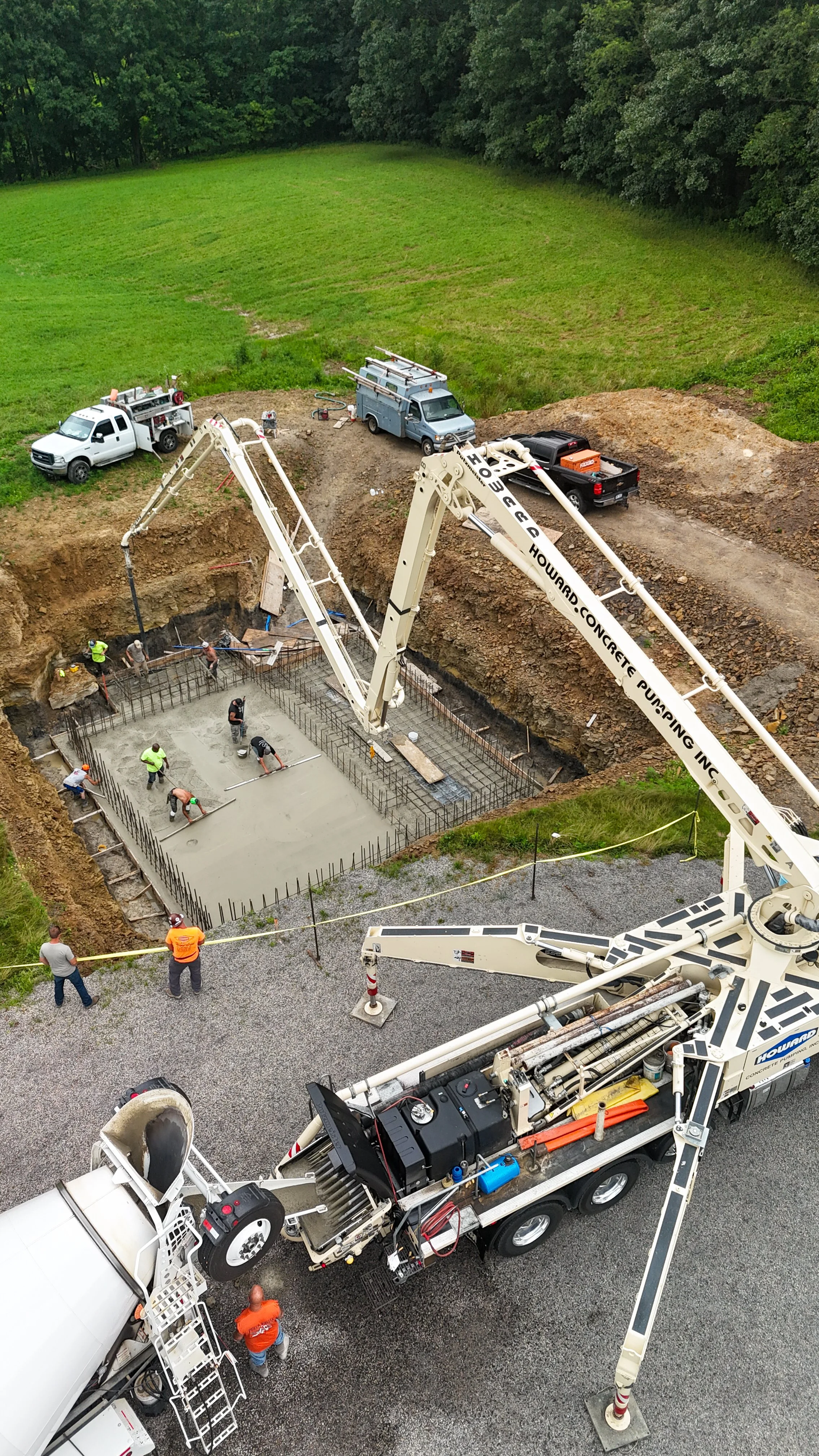 Construction workers are pouring a concrete foundation for a building, using a concrete pump truck and several small tools, with a green grassy field and trees in the background.