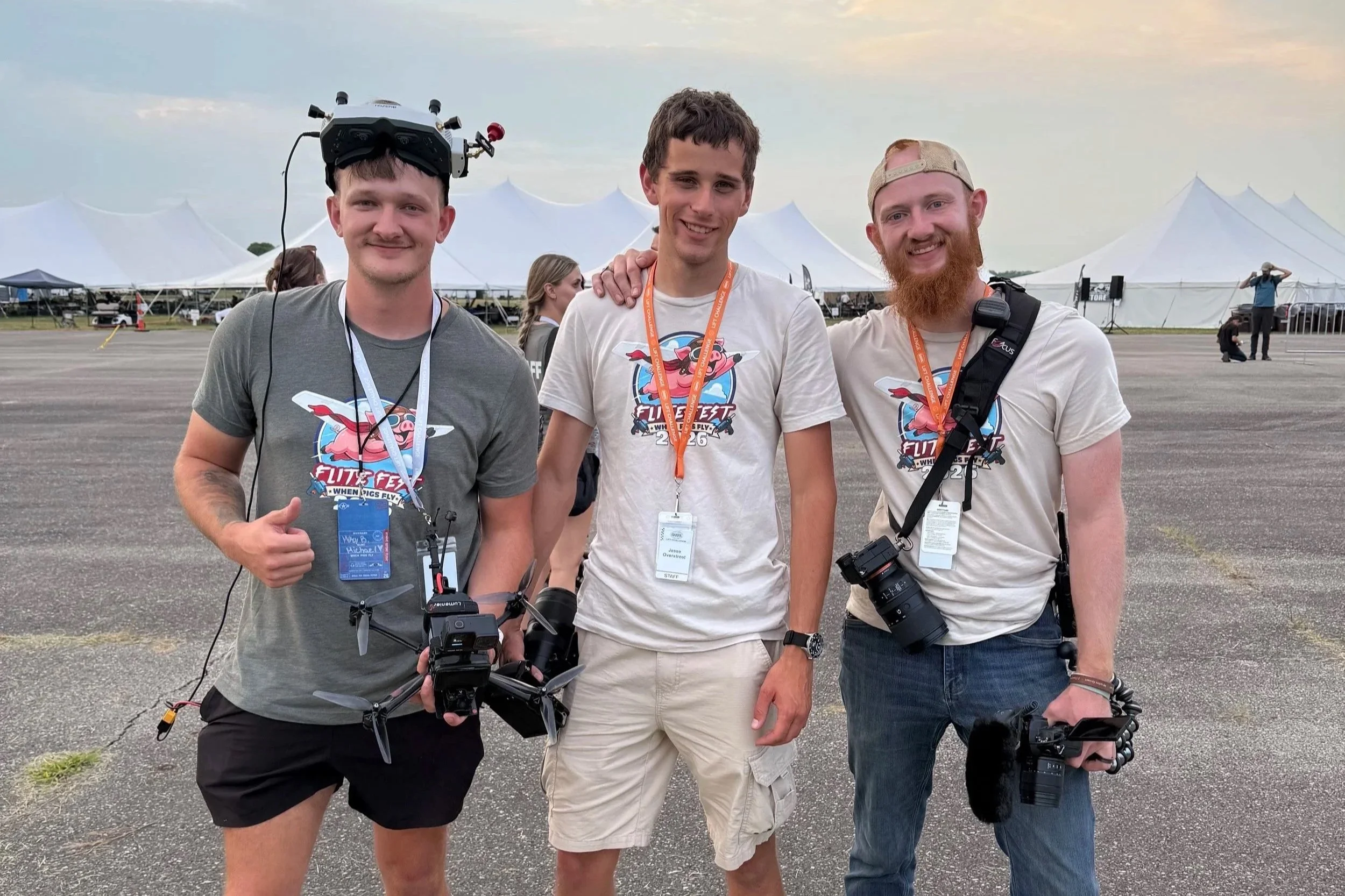 Three young men standing together at an outdoor event with large white tents in the background. All are smiling and wearing event badges and cameras. The person on the left is wearing a drone headgear, the middle person has his arm around the shoulder of the others, and the person on the right has a beard and a baseball cap worn backwards.