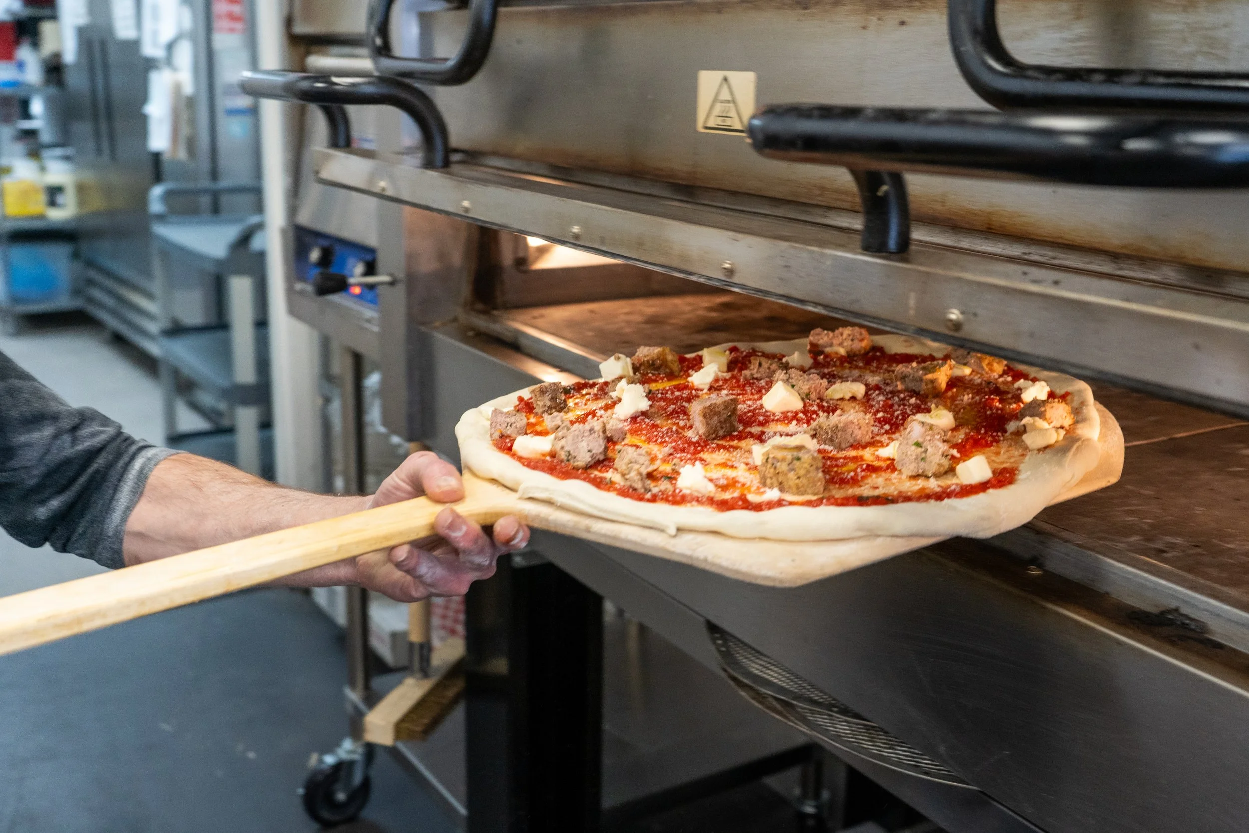 A person is placing a pizza into an oven. The pizza has toppings including cheese, sausage, and tomato sauce. The oven is a commercial pizza oven with a conveyor belt.