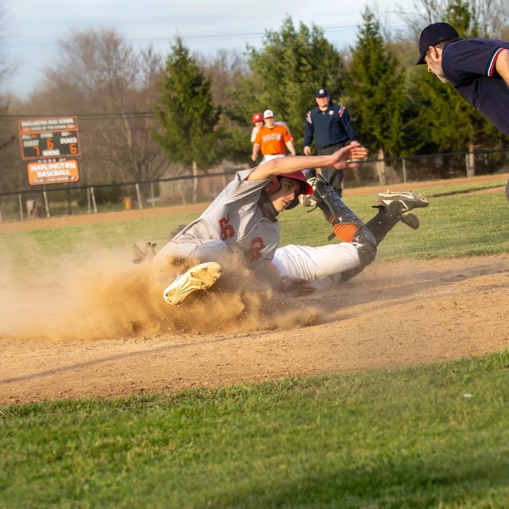 A baseball player in a gray uniform and red helmet slides into home plate while an umpire watches, with two players in orange and white uniforms and a man in a dark jacket in the background on a baseball field.