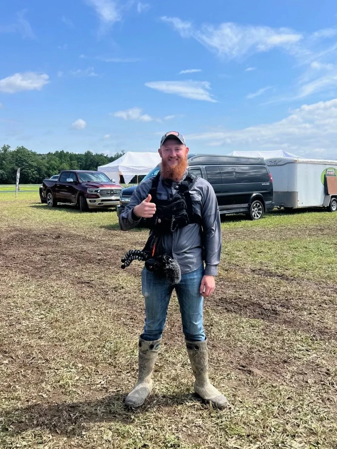 Man with a red beard wearing a gray jacket, jeans, and beige rubber boots standing outdoors on dirt ground, smiling and giving a thumbs-up. Behind him are several parked trucks and a trailer under a partly cloudy sky.