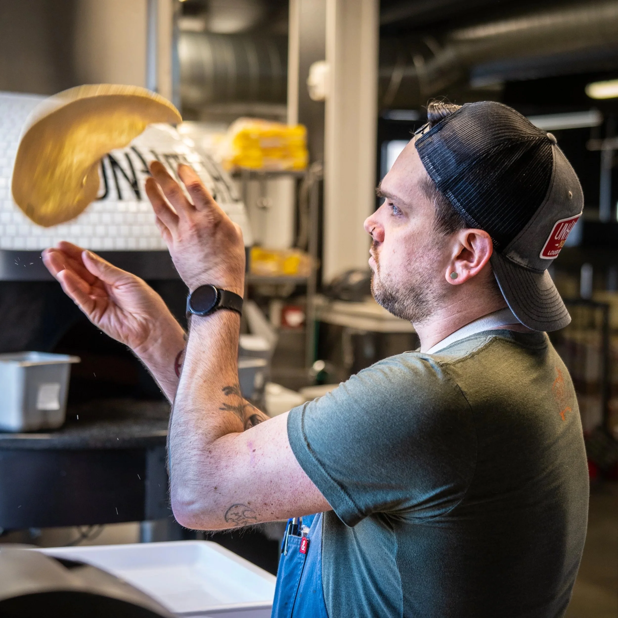 A person with a tattoo on their arm and piercings in their ear, wearing a black cap, blue jeans, and a gray hoodie, is reaching up towards shelves in what appears to be a commercial kitchen or storage area.