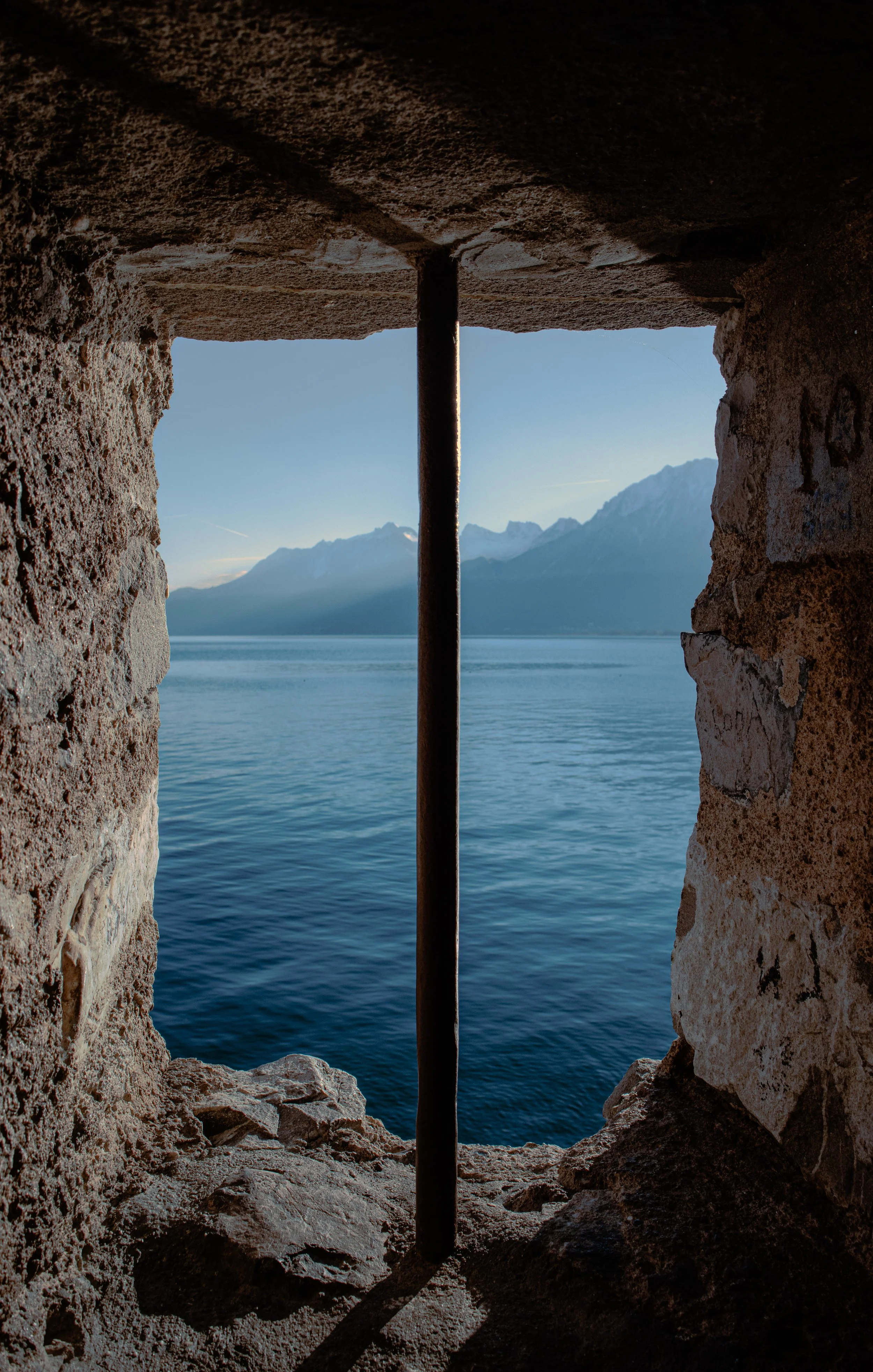 View of a lake and mountains through a small, rectangular opening in a rocky wall with a metal bar in the center.