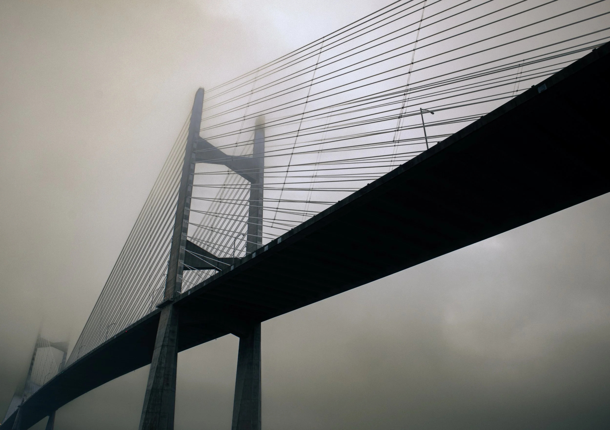 A modern cable-stayed bridge with multiple cables extending from tall, angled towers, seen partly obscured by fog or mist.