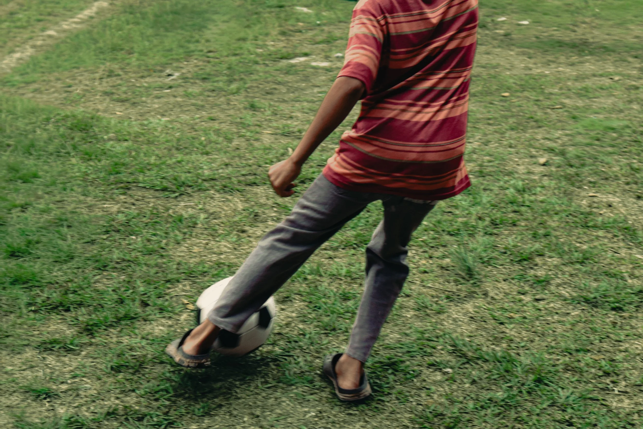 Person wearing a red and orange striped shirt and gray pants playing soccer on a grassy field.