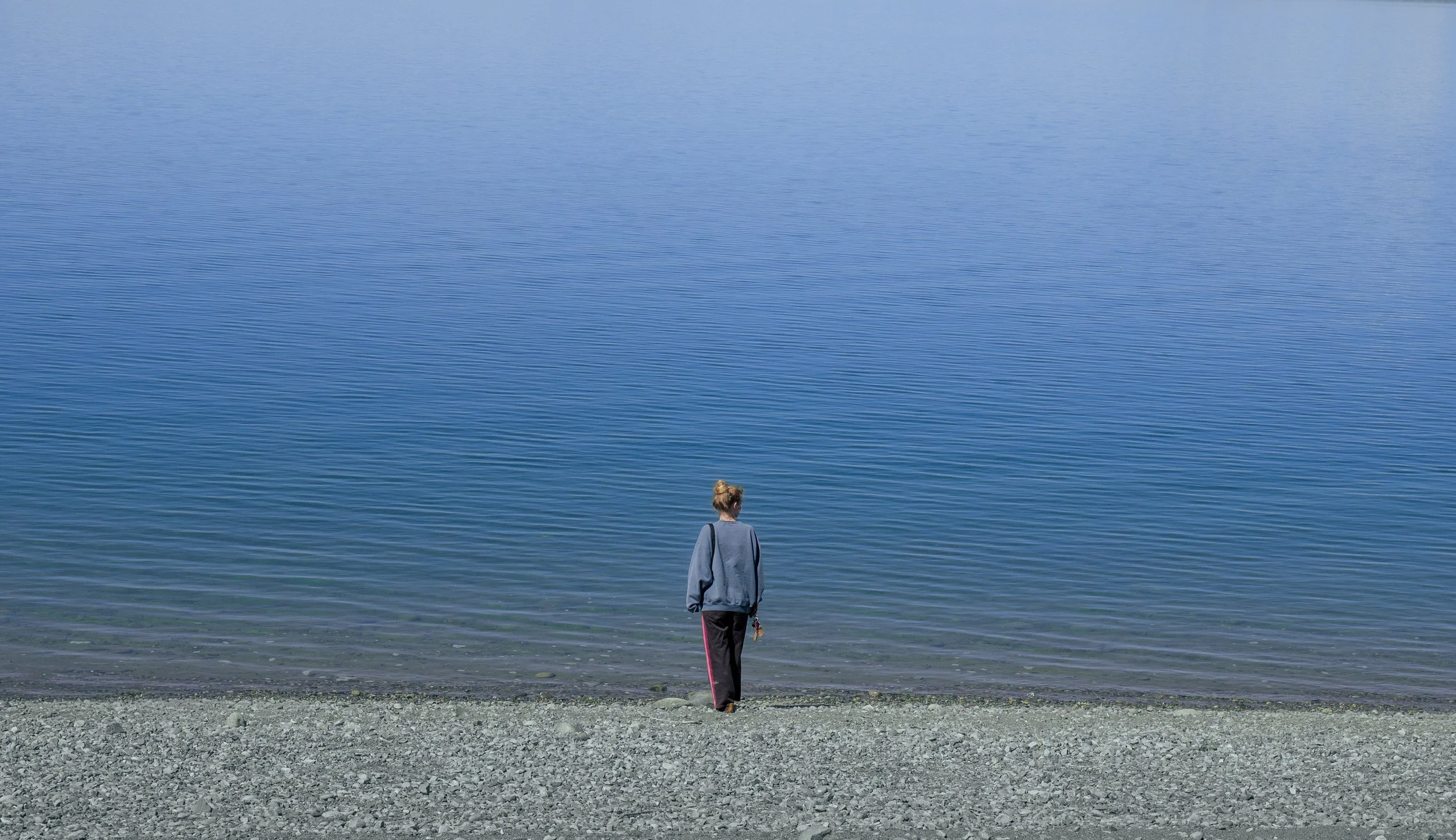 A person with blonde hair in a bun, wearing a gray jacket, black pants, and holding a small item, standing on a gravelly shore and looking at blue water.