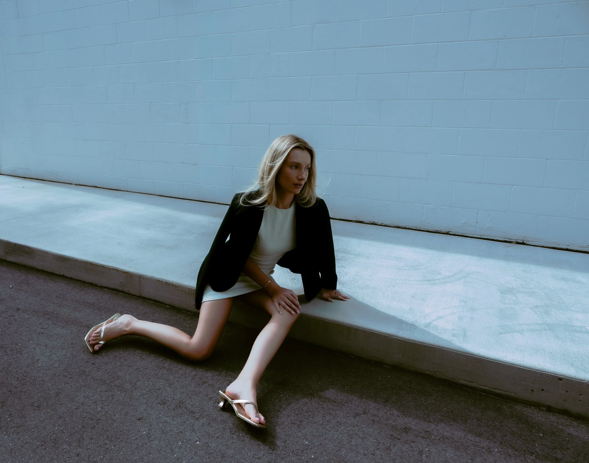Young woman sitting on the ground outside, next to a light blue brick wall, wearing a black blazer, white top, and white sandals with heels, with a serious facial expression.