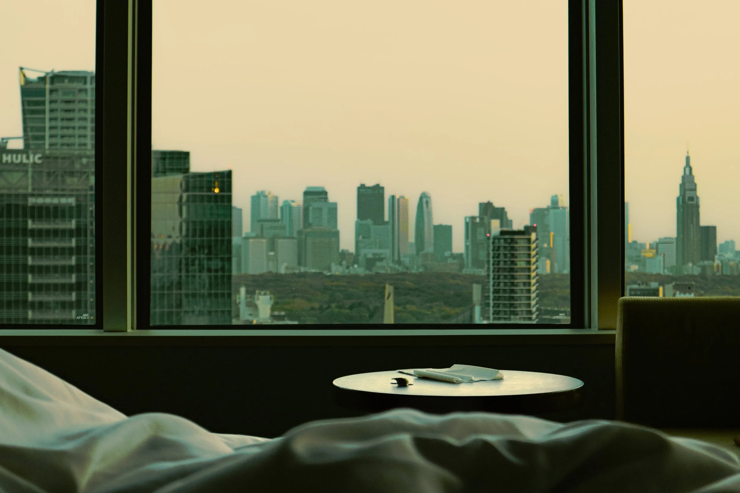 View from a hotel room window showing a city skyline with tall buildings and skyscrapers at sunset, with partially visible bed in the foreground.